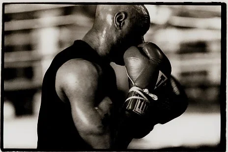 A boxer in a black sleeveless shirt with gloves, standing in a boxing stance with head bowed and fists up.