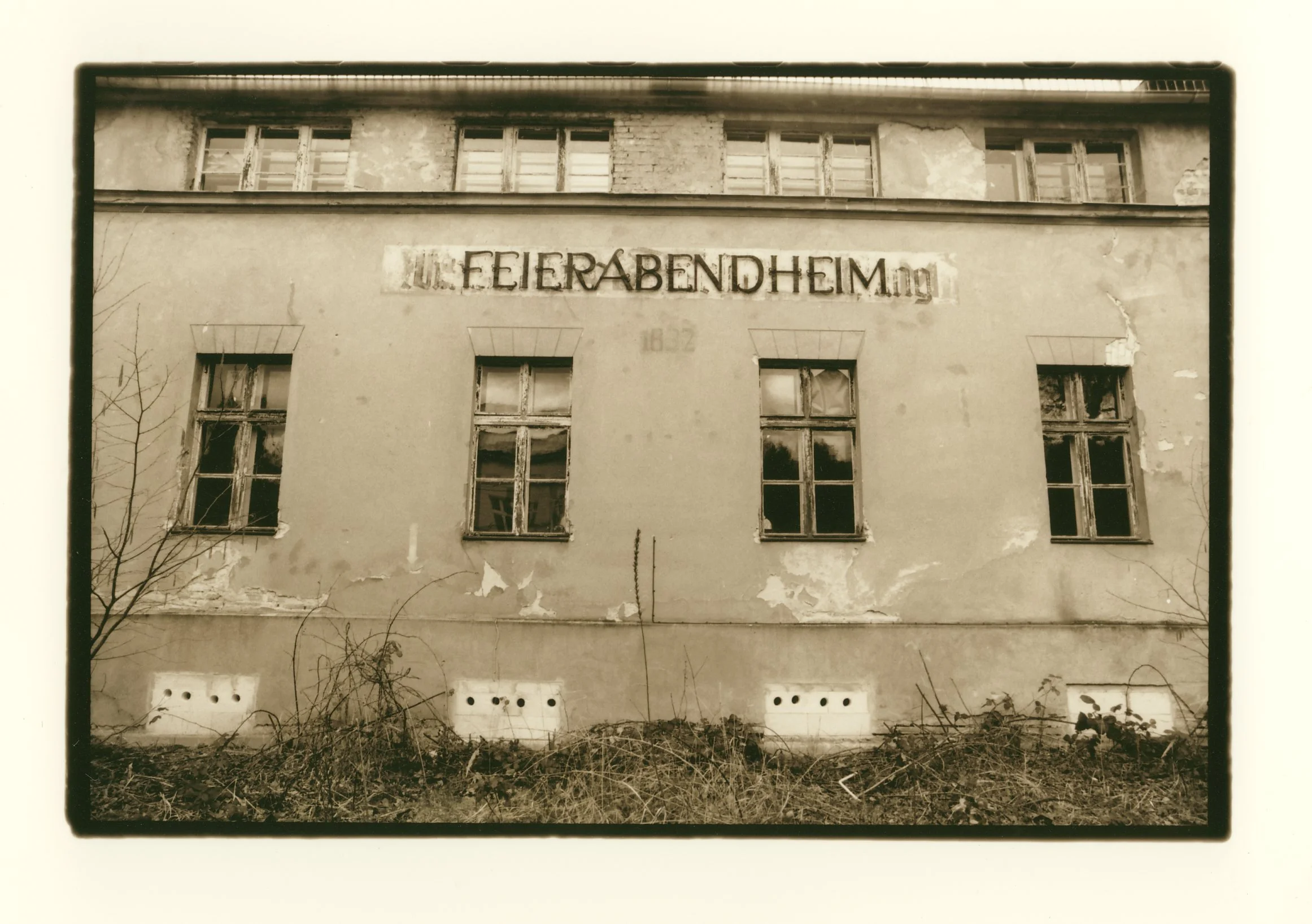 An old, neglected building with peeling paint and boarded-up windows, with a faded sign reading 'Feierabendheim' on the wall, in sepia tone.