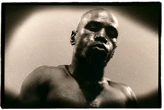 Black and white photo of a shirtless man with a serious expression, looking directly at the camera.