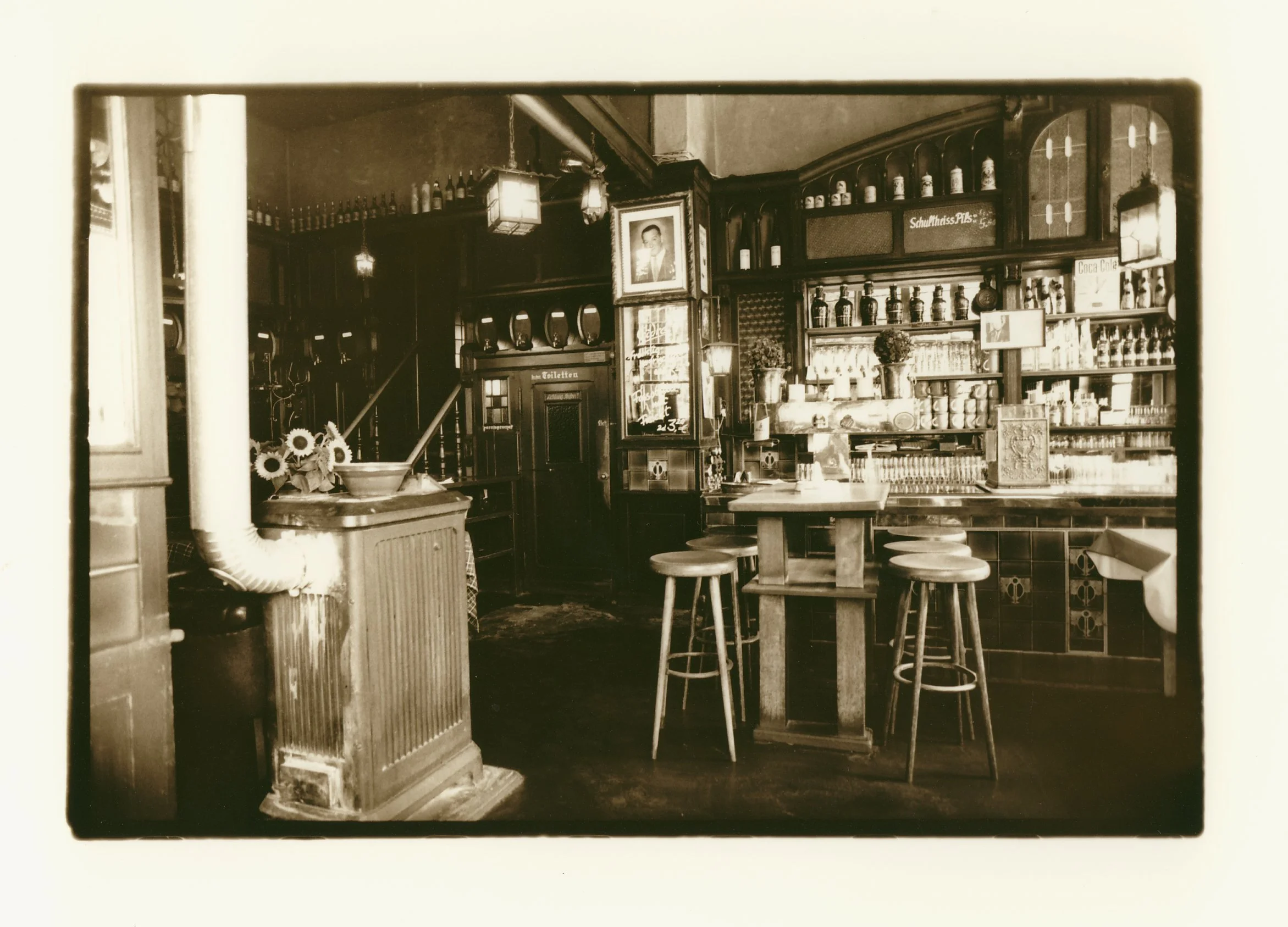 A vintage black-and-white photograph of a cozy, old-fashioned café or bar interior with wooden furniture, shelves filled with bottles and jars, framed photographs on the wall, and a counter with stools.