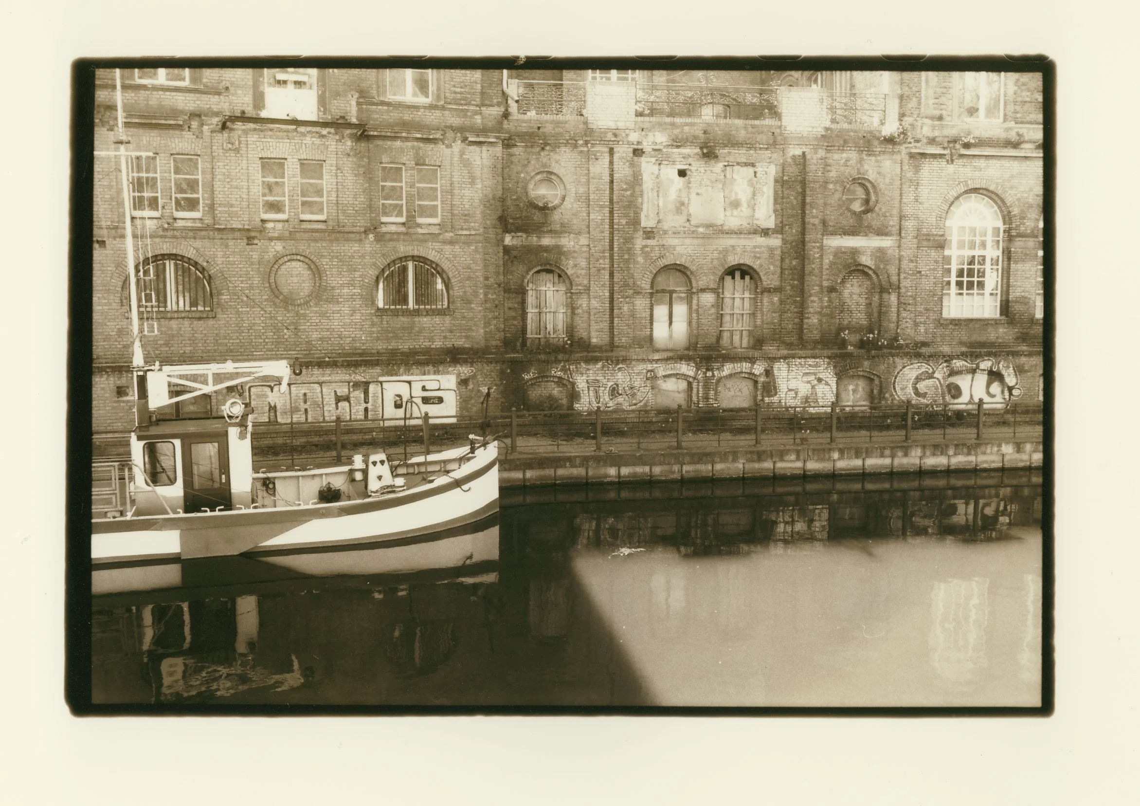 Sepia-toned photograph of a boat docked along a canal with a brick building covered in graffiti in the background.