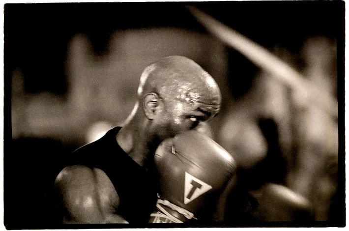A male boxer with a shaved head wearing boxing gloves and in a fighting stance in a boxing ring.