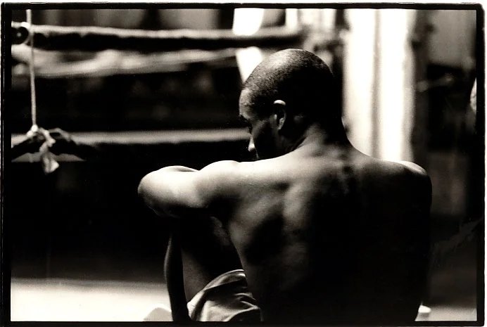 A black and white photo of a shirtless man sitting with his back to the camera, resting his chin on his knee in a dimly lit room.