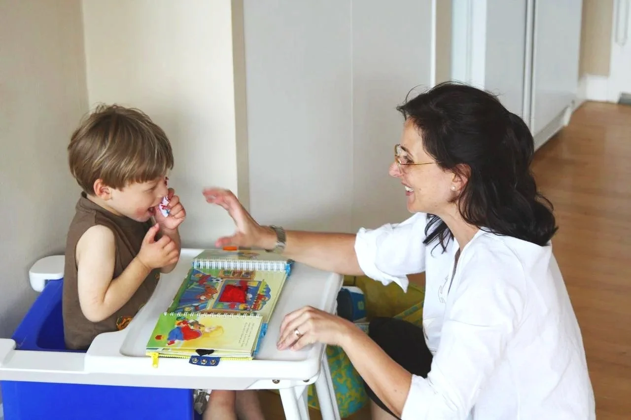 Niño pequeño en silla comiendo, mujer sonriendo y alcanzandolo, en una habitación interior