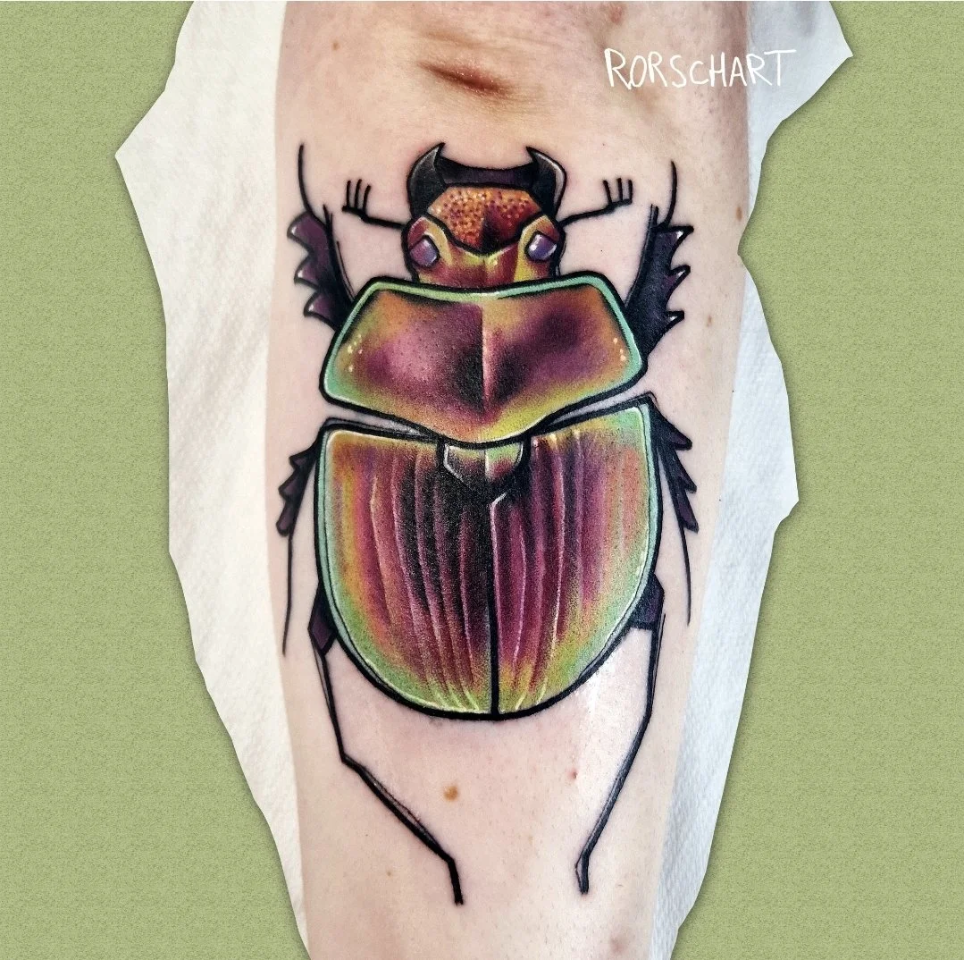 L'image montre un tatouage représentant un scarabée semi réaliste dans une gamme de couleurs violette, jaune et vert. Le style graphique est contemporain, légèrement déstructuré et très coloré. Le tatouage a été réalisé par Rorschart, tatoueur à Nanc