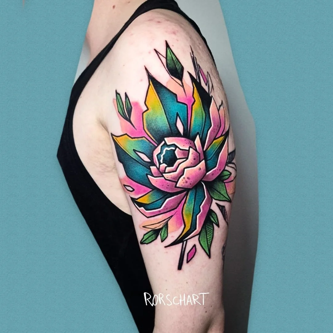 L'image montre un tatouage d'une fleur sur une épaule. Le style graphique est contemporain, légèrement déstructuré et très coloré. Le tatouage a été réalisé par Rorschart, tatoueur à Nancy.