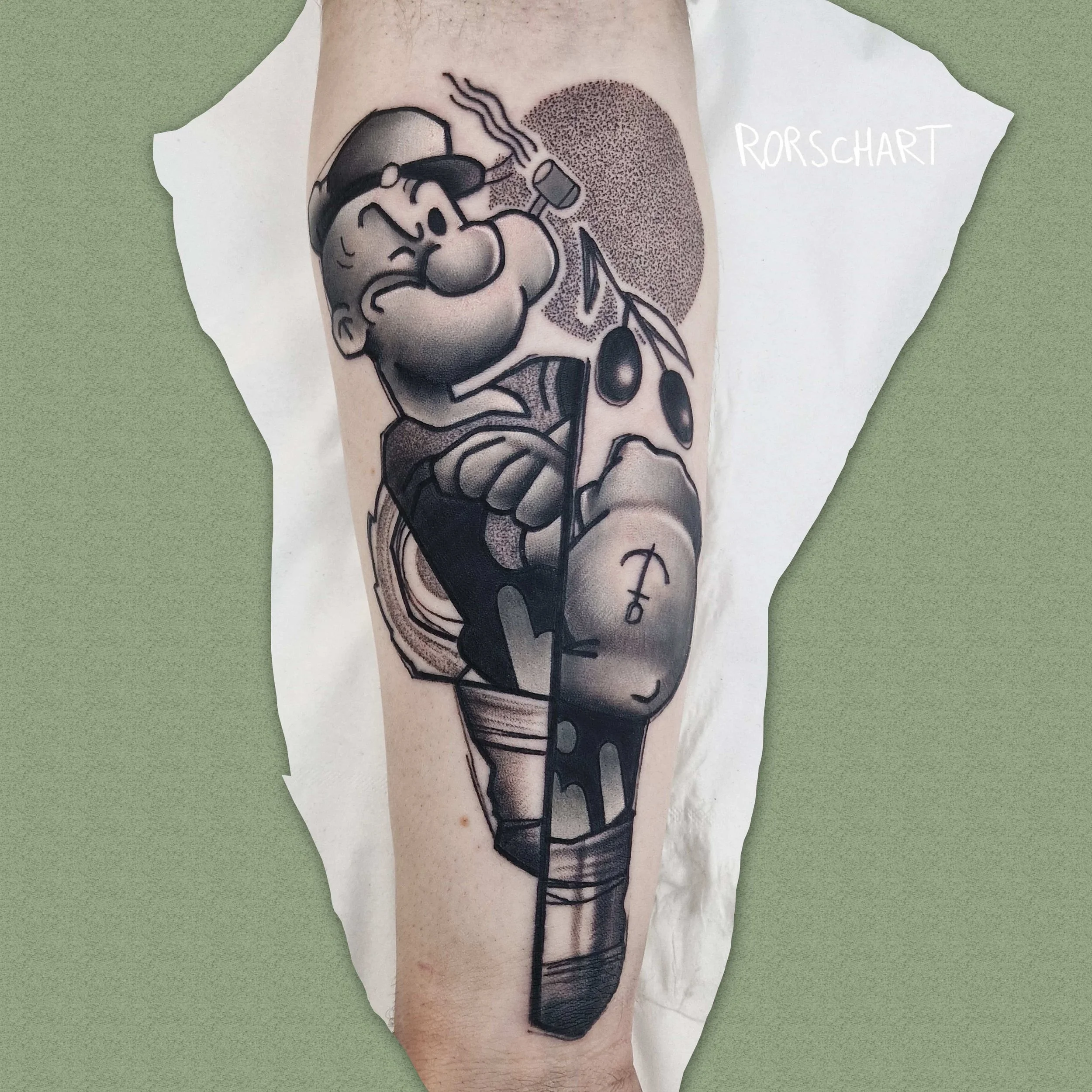 L'image montre un tatouage représentant Popeye avec une boîte de conserve et une branche d'olivier sur un avant bras. Le style graphique est contemporain, légèrement déstructuré et en noir et gris. Le tatouage a été réalisé par Rorschart, tatoueur à