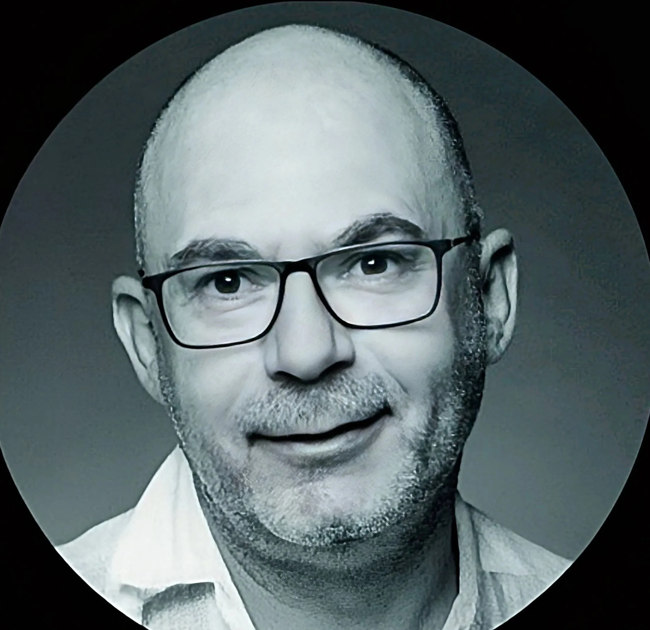 Portrait d'un homme portant des lunettes, avec une barbe grise, souriant légèrement, en noir et blanc.