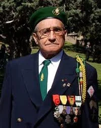 Un homme âgé en uniforme militaire ou de service, portant des décorations et des médailles, avec un béret vert, à l'extérieur, probablement lors d'une cérémonie ou d'un événement commémoratif.