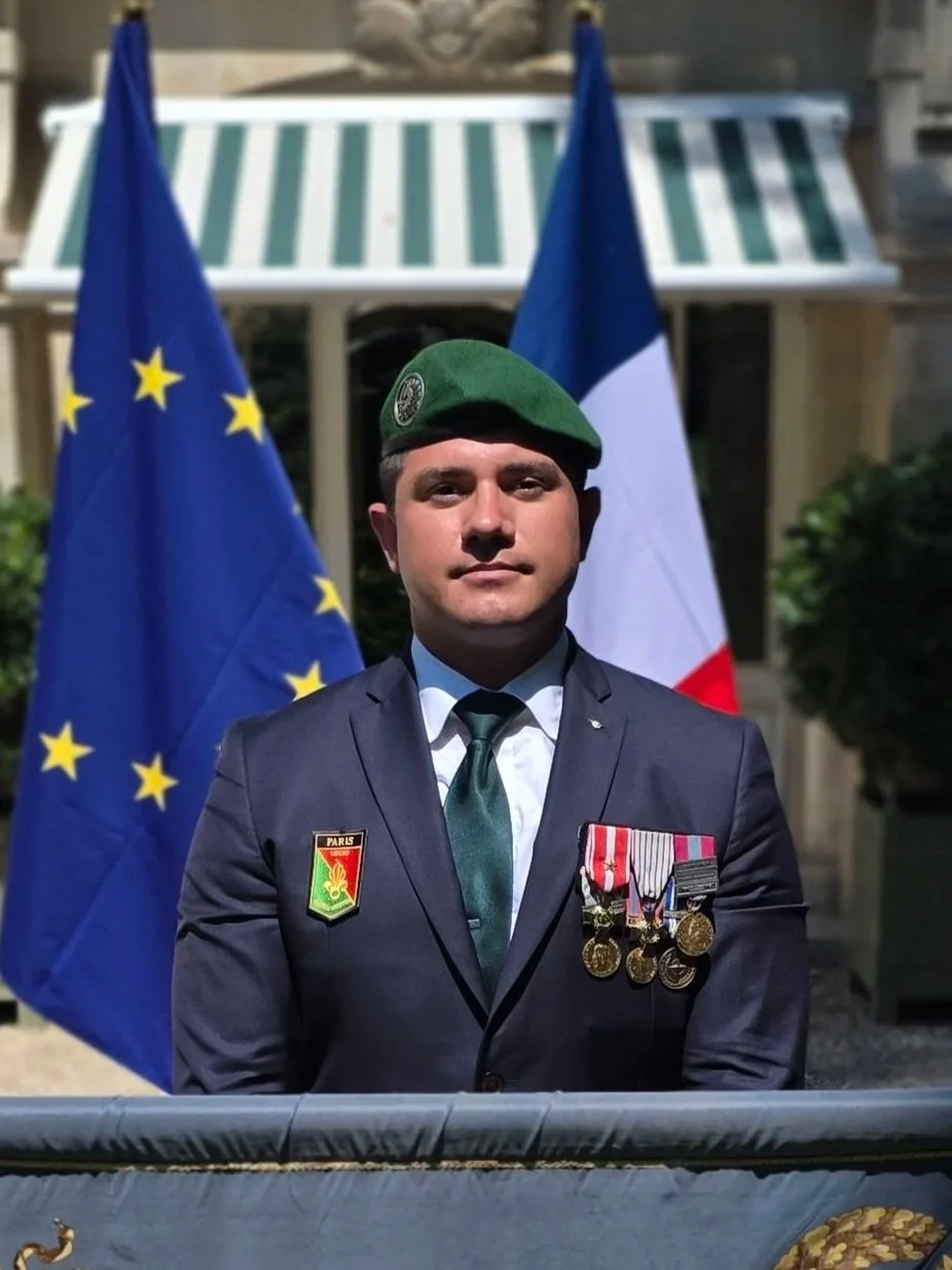 Un homme en uniforme militaire avec des médailles, portant un béret vert, se tenant devant les drapeaux de l'Union européenne et de la France.