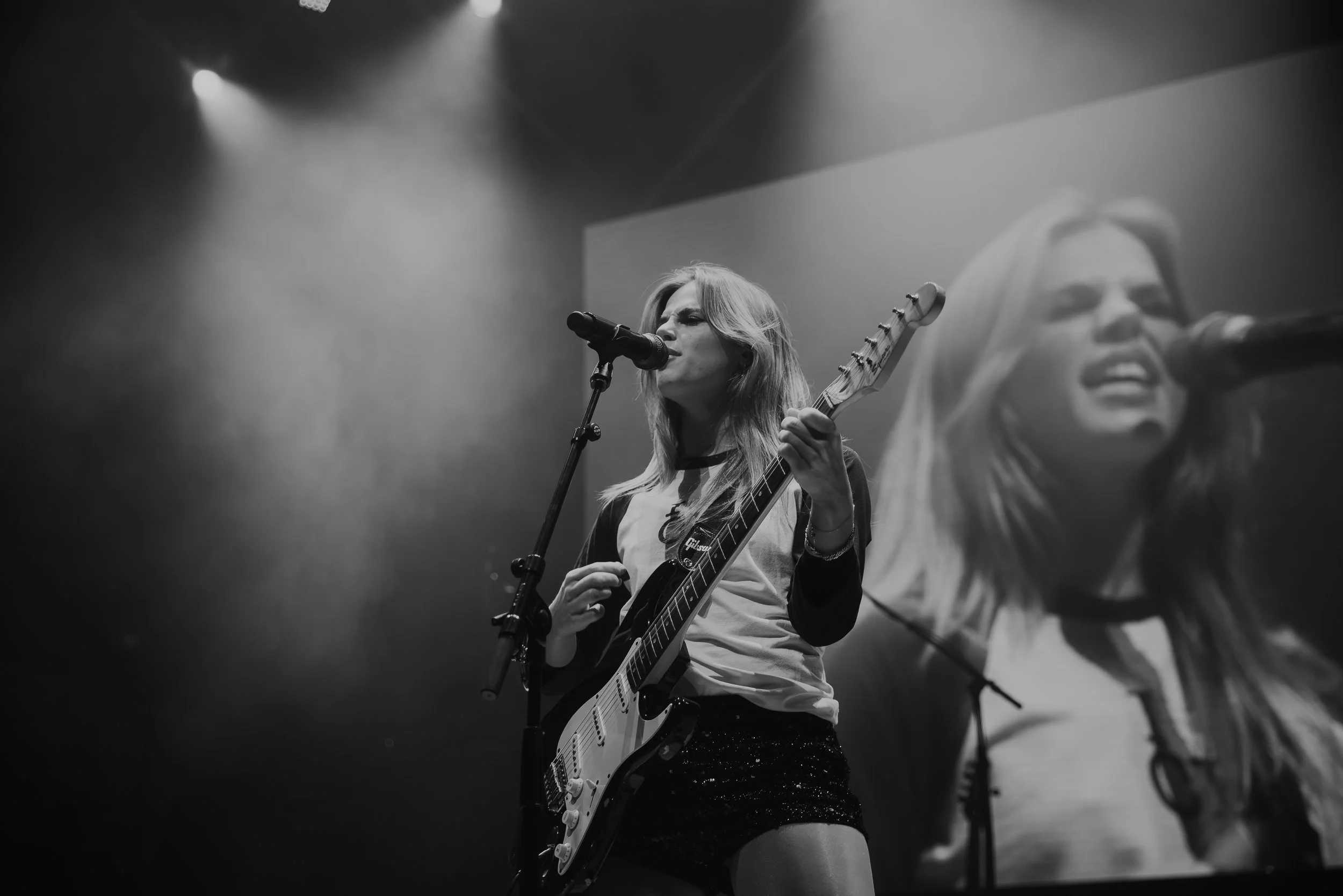 Cassie Henderson singing into a microphone while playing an electric guitar on stage, with a large screen behind her showing her face.
