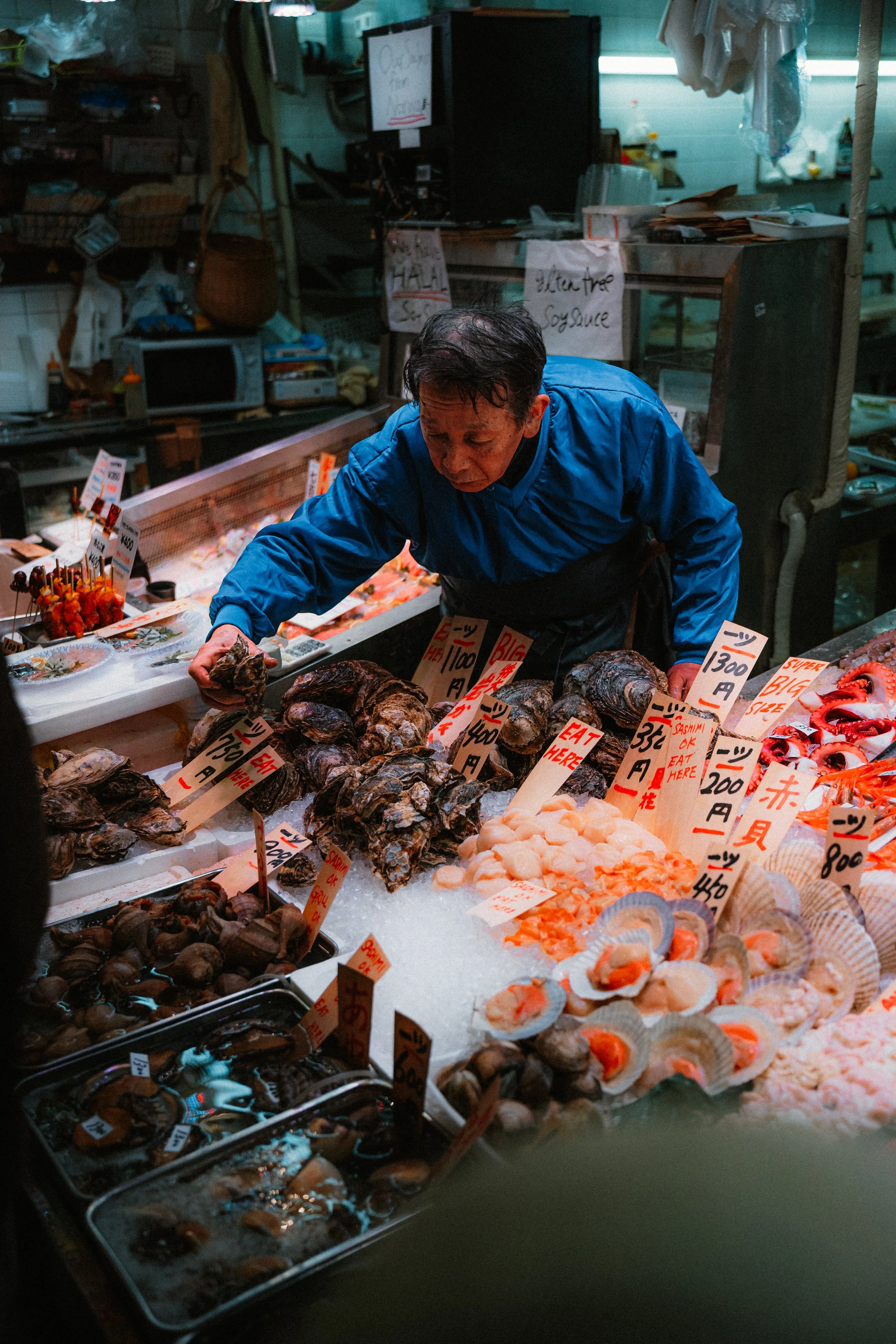 A man in a blue jacket working at a seafood market stall. The stall has various shellfish and seafood displayed on ice, with signs and price tags in Japanese and English.