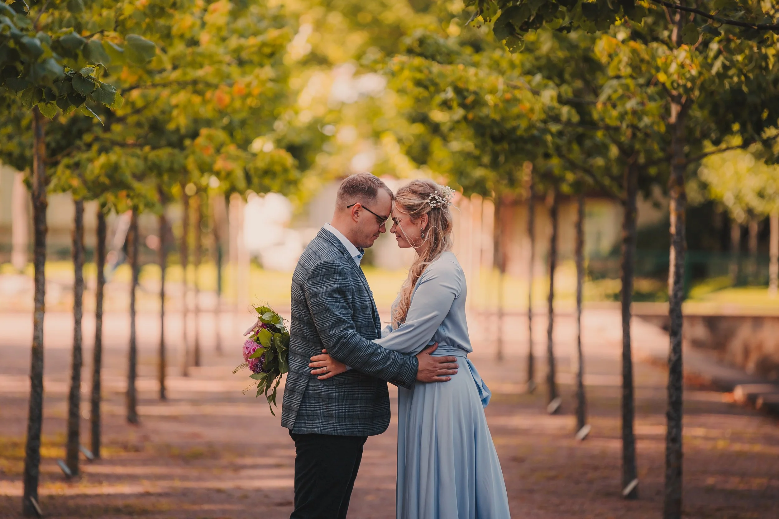 A couple, dressed in formal wedding attire, holding each other with foreheads touching in an outdoor park setting with green trees and sunlight.