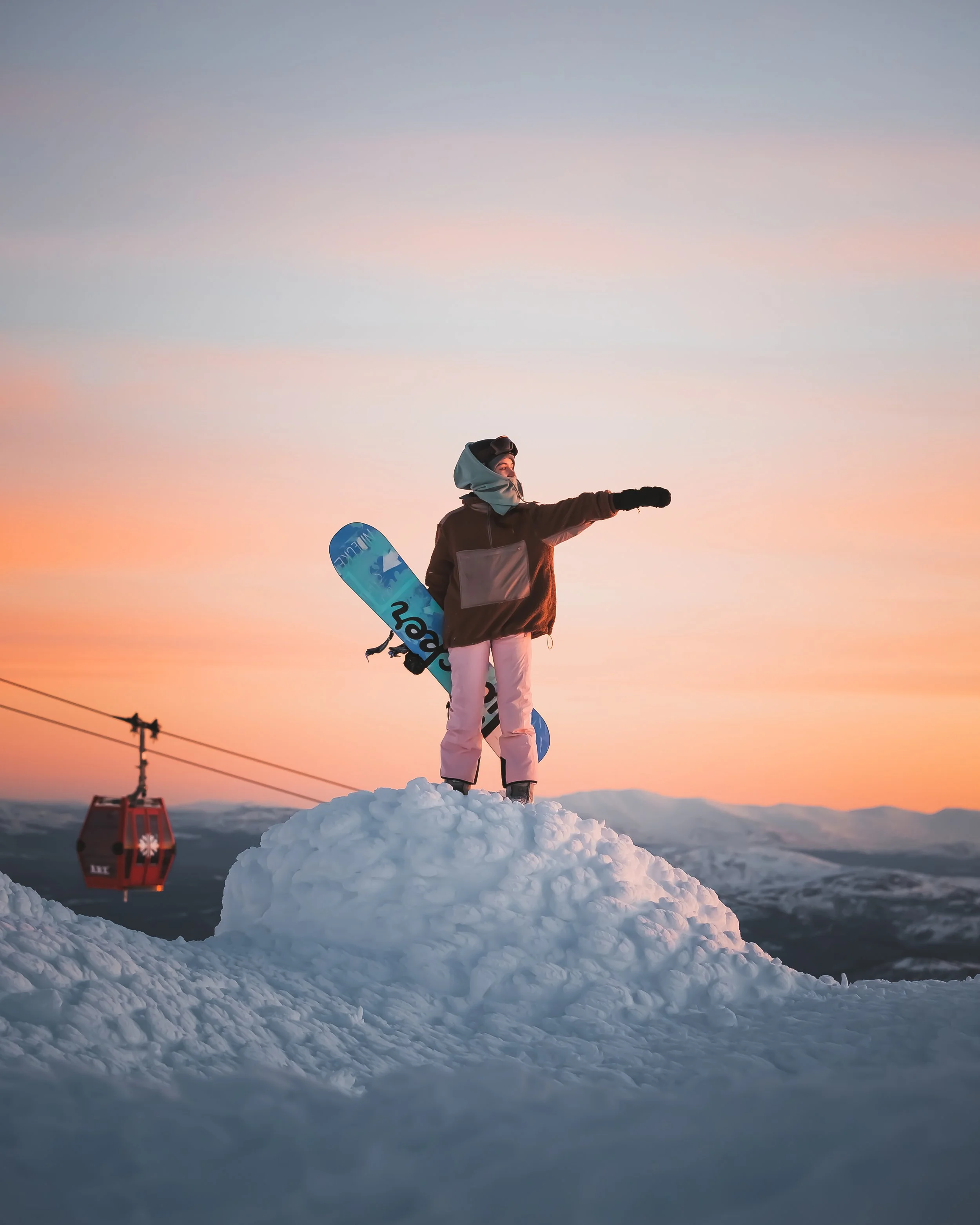 A person dressed in winter clothing, standing on a snow mound at sunset, holding a snowboard, with a ski lift in the background.