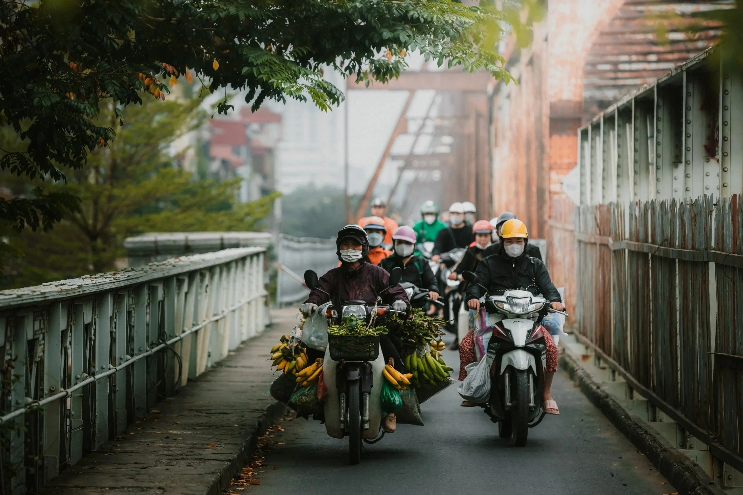 Group of people riding motorcycles and scooters on a narrow urban street, wearing helmets and face masks, with one motorcycle loaded with bananas and vegetables, in a city setting with trees and brick buildings.