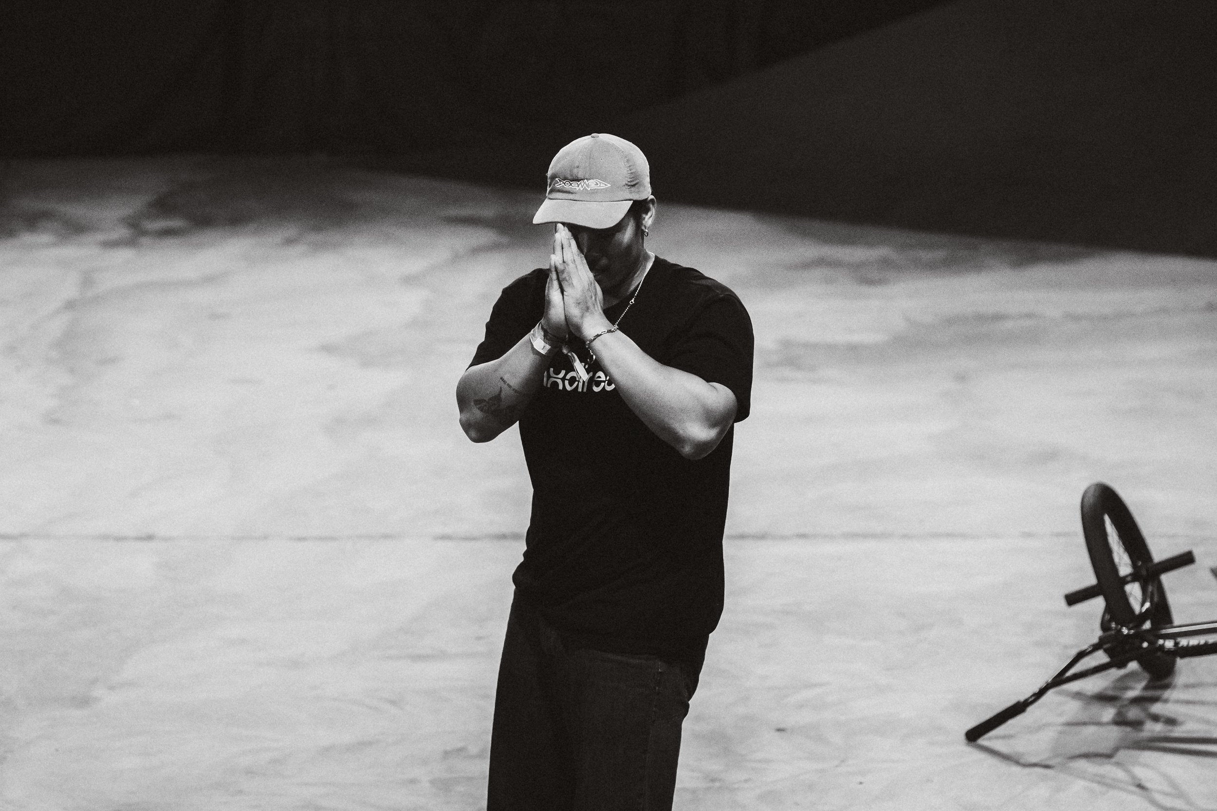 A man wearing a cap and a black t-shirt stands on a stage with his hands pressed together in prayer or gratitude. A bike is lying on the ground beside him.