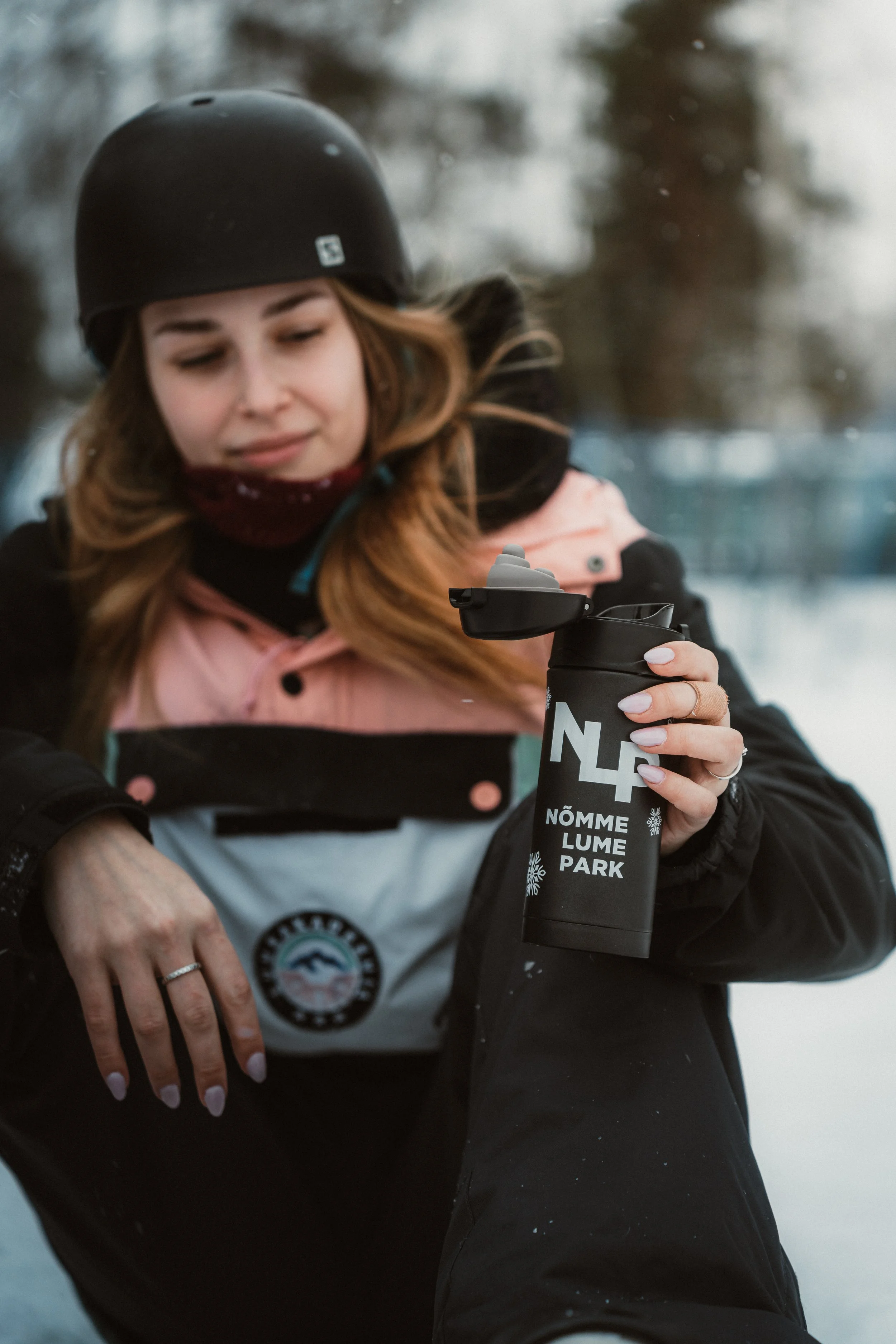 A woman in winter gear holding a black water bottle with the text "NÕMME LUMEPARK" during winter.