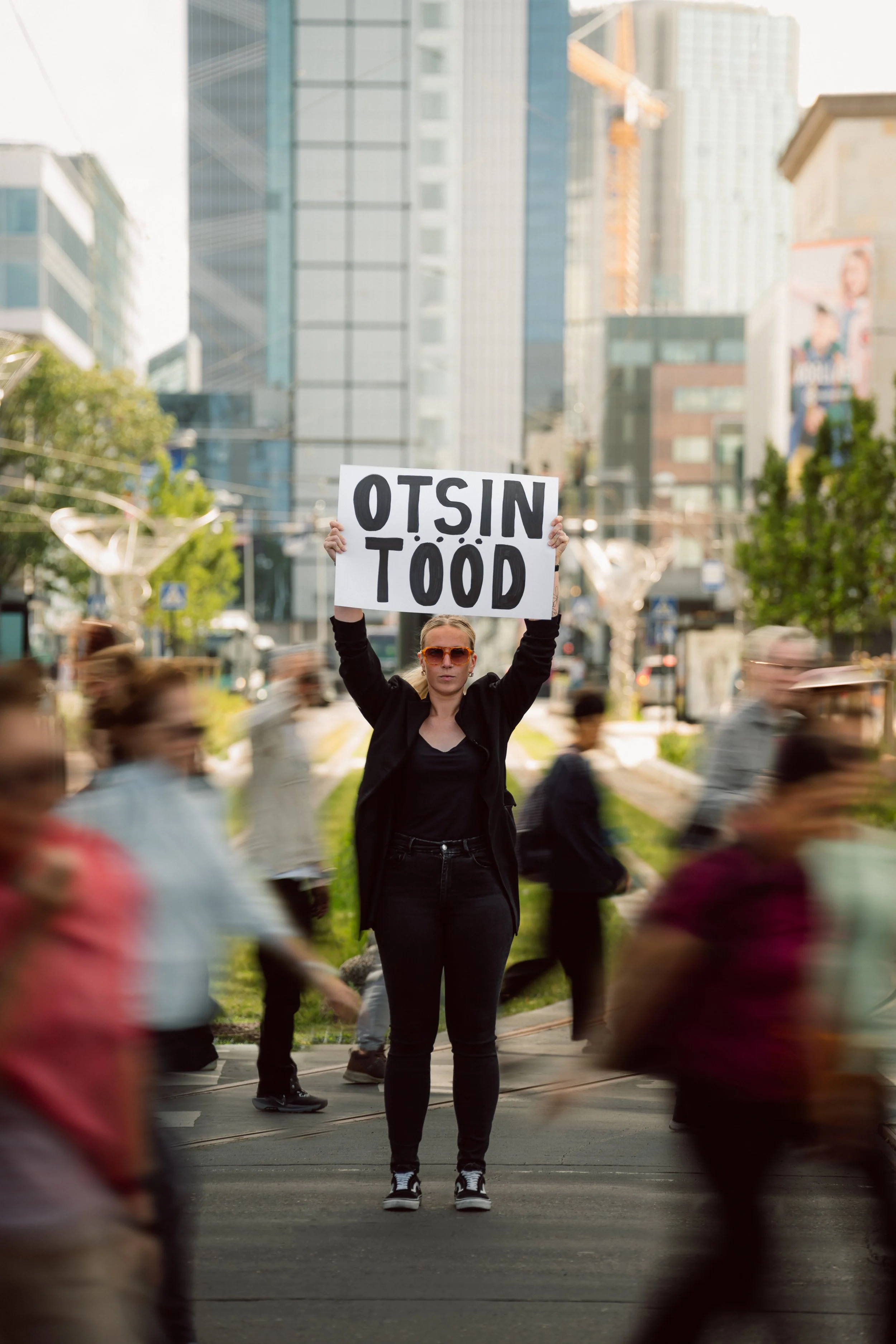 A woman standing in an urban street  holding a sign that says 'OTSIN TÖÖD', with blurred pedestrians walking past and tall buildings in the background.
