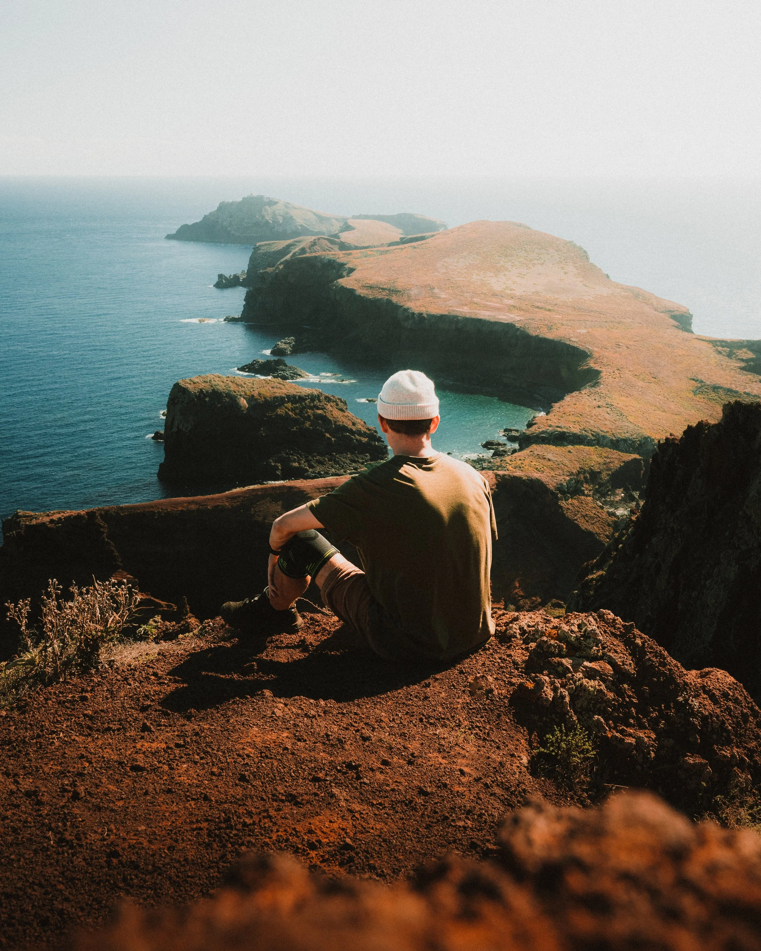 A person wearing a white beanie and dark clothing, sitting on a rocky cliff overlooking a coastal landscape with cliffs and ocean in the distance.