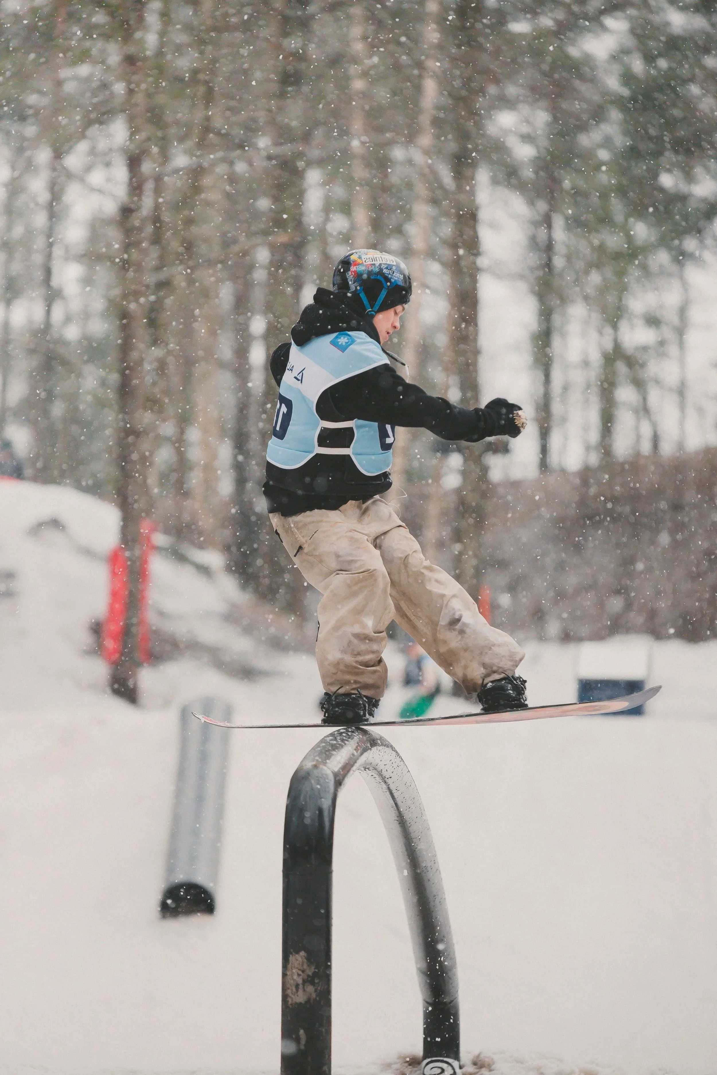 A young snowboarder in a blue helmet and jacket performs a trick on a rail at a snowy outdoor skate park.