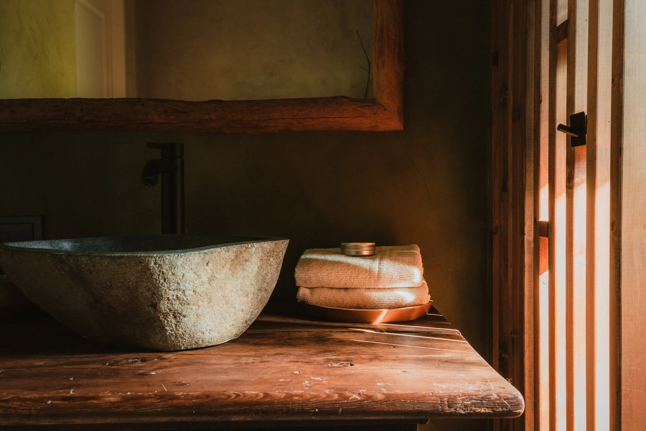 Close-up of a rustic wooden bathroom vanity with a stone vessel sink, a black faucet, and neatly folded towels with a small container on top. Warm lighting filters through wooden slats on the side.
