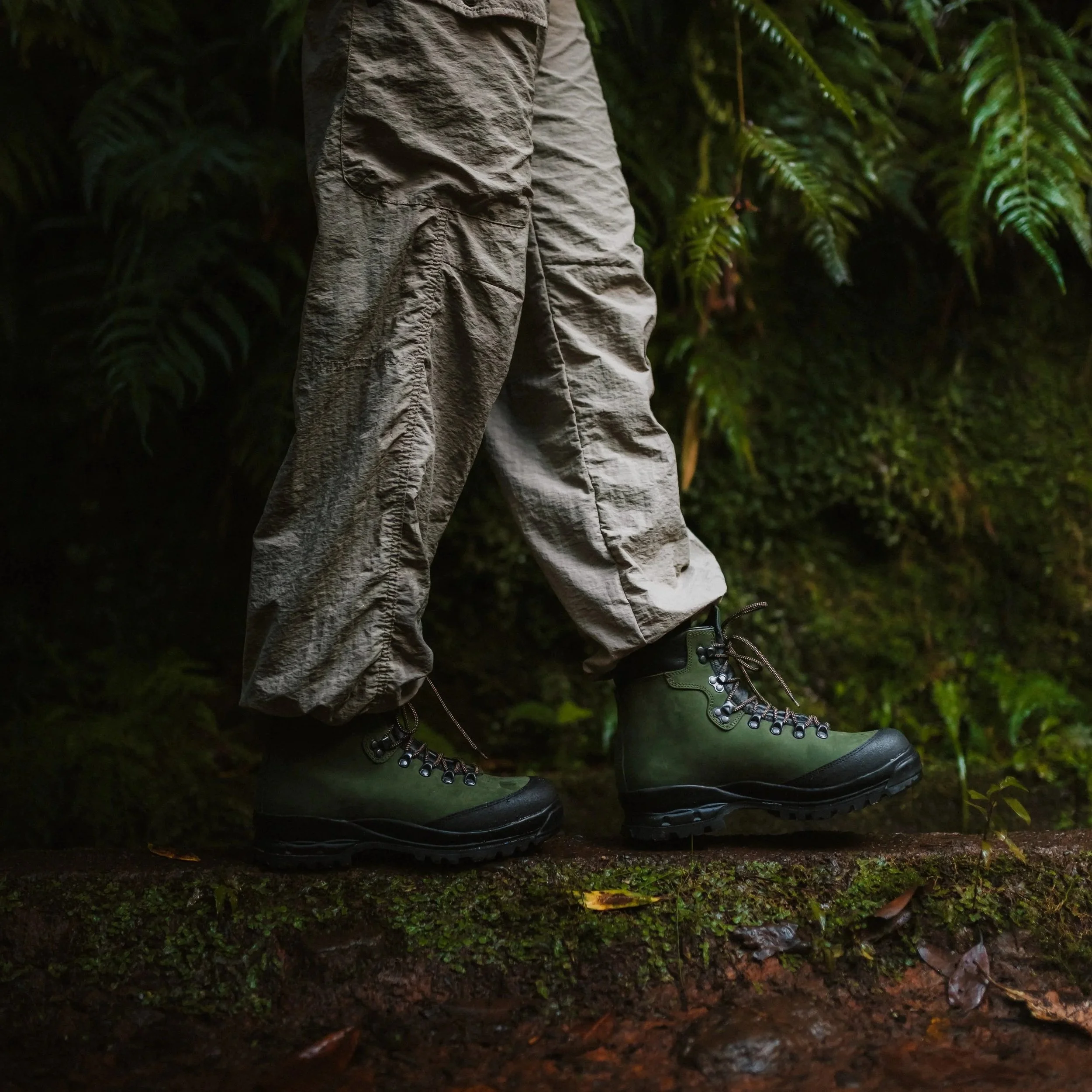 Close-up of a person wearing green hiking boots and khaki cargo pants standing on a mossy and leaf-covered trail in a lush forest.