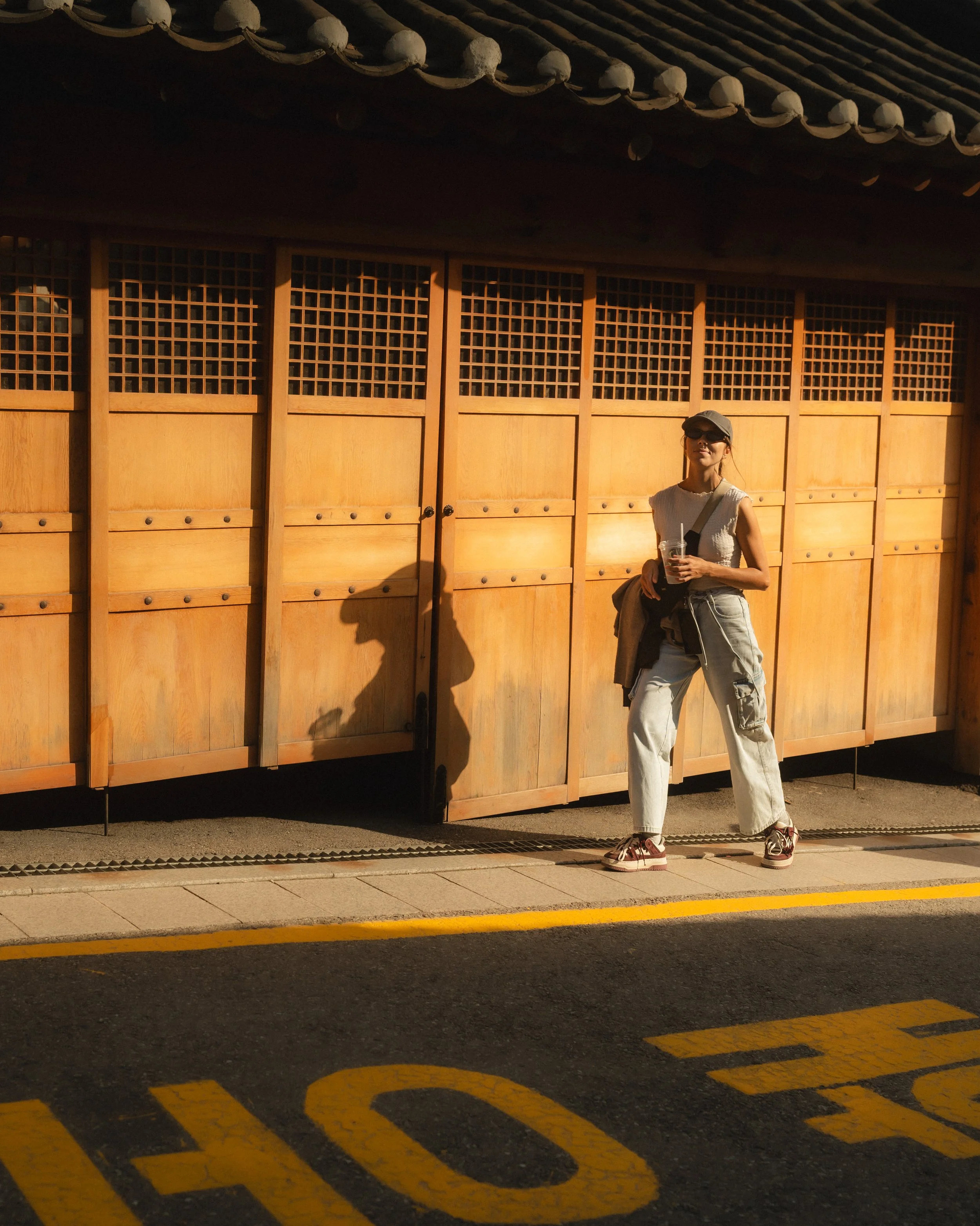 A woman standing outside a traditional wooden structure, wearing sunglasses, a sleeveless top, cargo pants, and sneakers, holding a drink, with her shadow cast on the wooden wall.