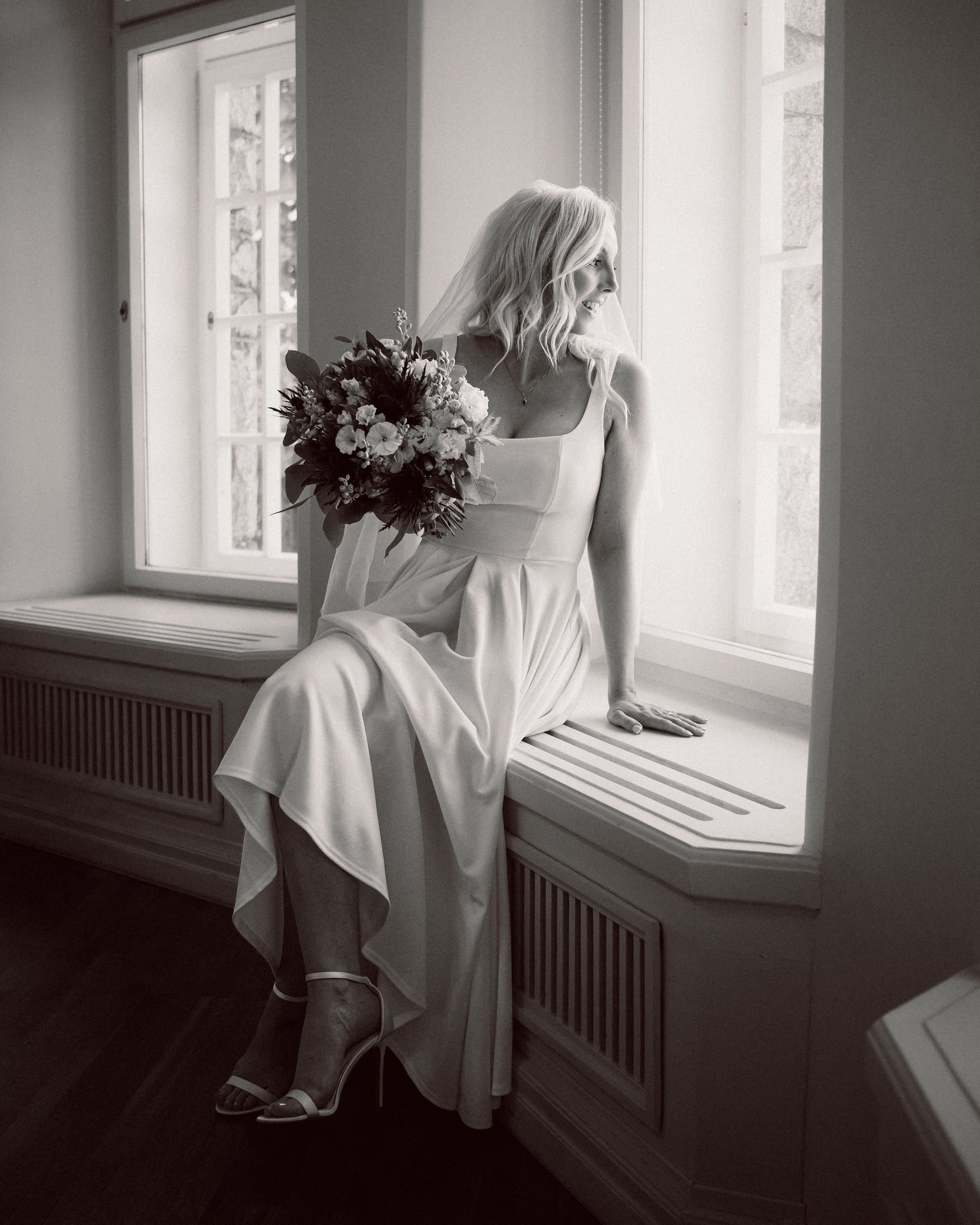 Black and white photo of a bride sitting on a window seat, holding a bouquet, dressed in a wedding gown, looking out the window.
