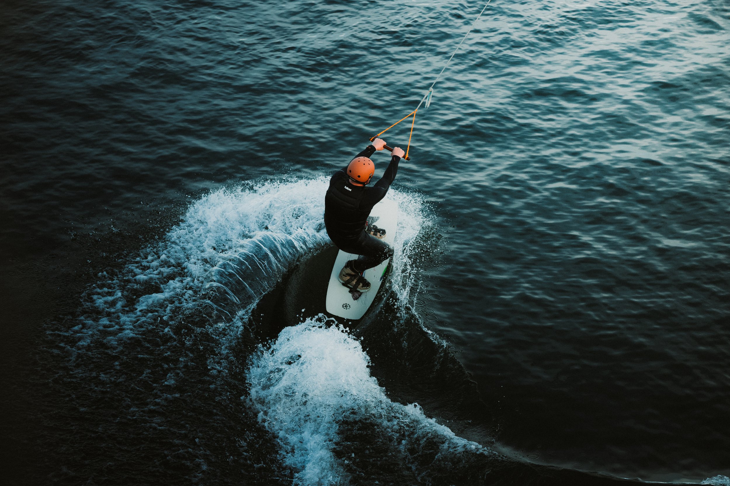 A person wearing a black wetsuit and an orange helmet rides a wakeboard on a boat, holding a handle attached to a cable, as they carve through the water.