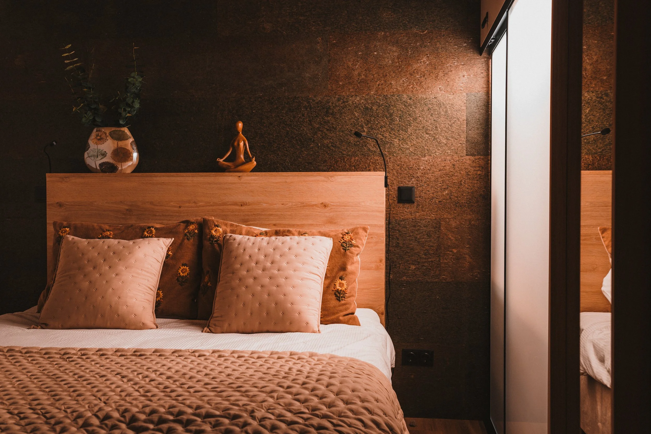 Cozy bedroom with wooden headboard, decorative pillows, and an amber quilt. A dark textured wall features a vase with green foliage and a wooden figurine. A mirrored wardrobe reflects part of the bed, with nearby wall-mounted lamps and electrical switch.