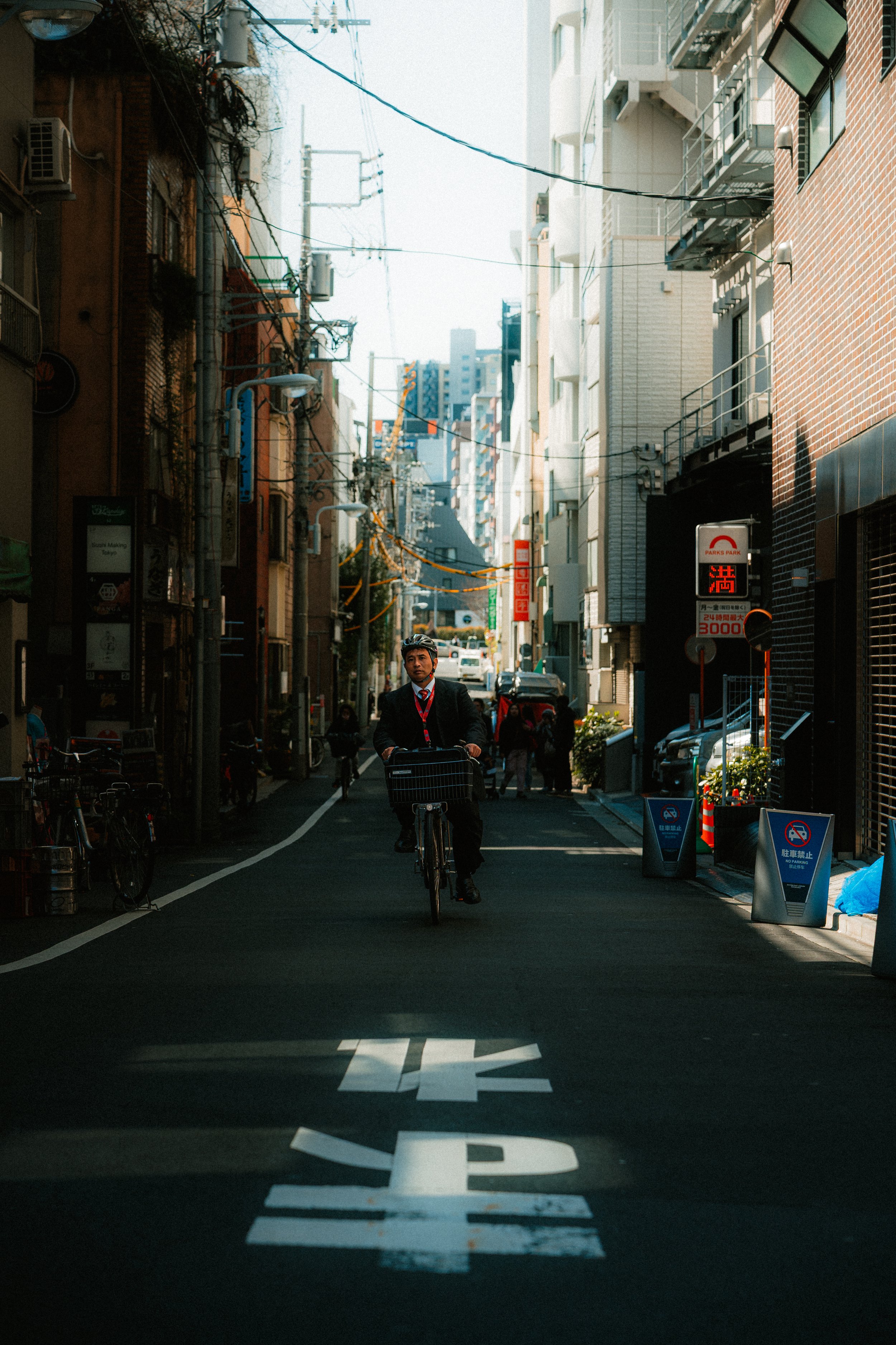 A man in a business suit riding a bicycle on a city street with tall buildings, parked cars, and pedestrians in the background.