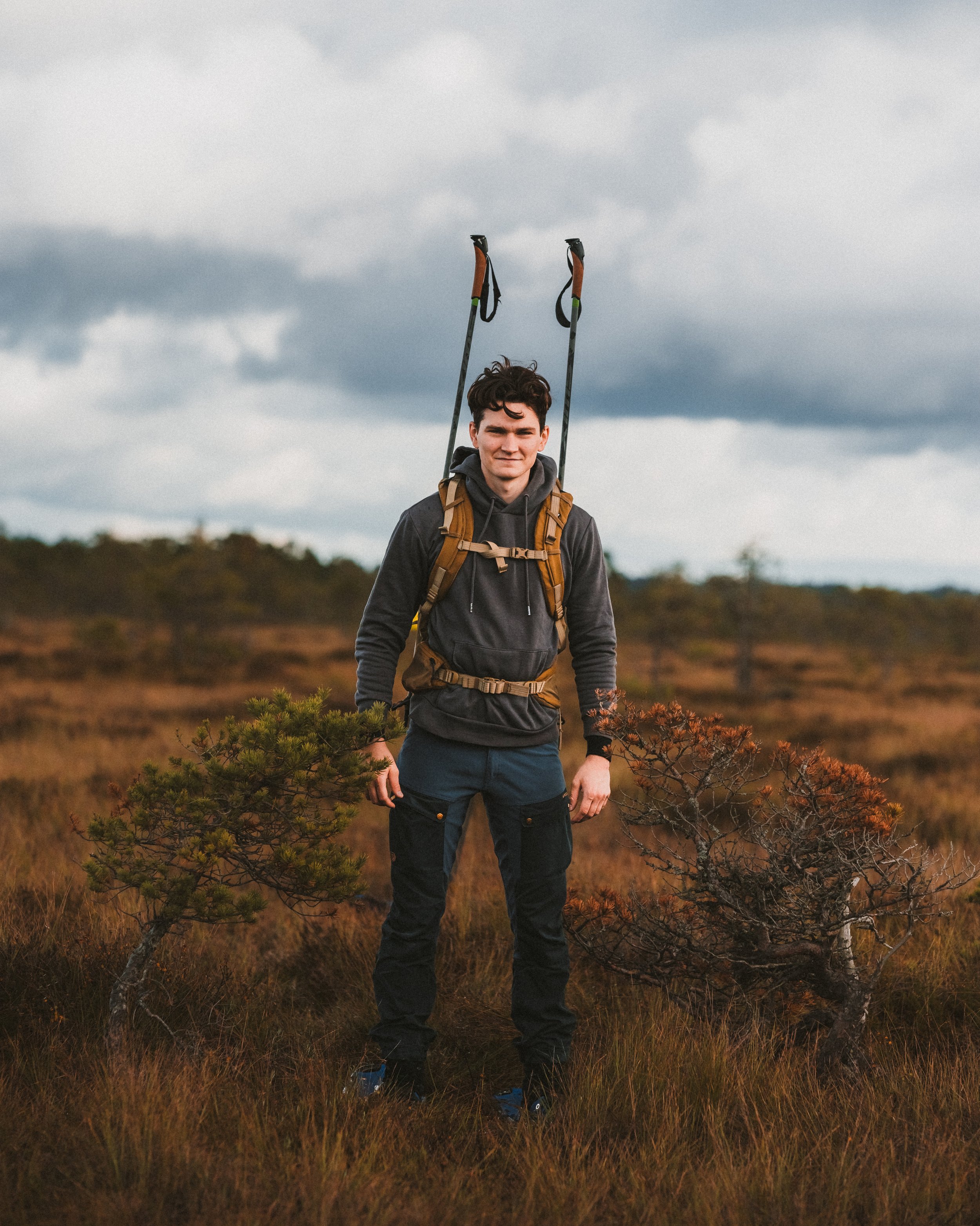Young man standing outdoors in a grassy field with small trees, wearing a black hoodie, dark pants, a backpack with hiking poles attached, and looking at the camera under cloudy skies.