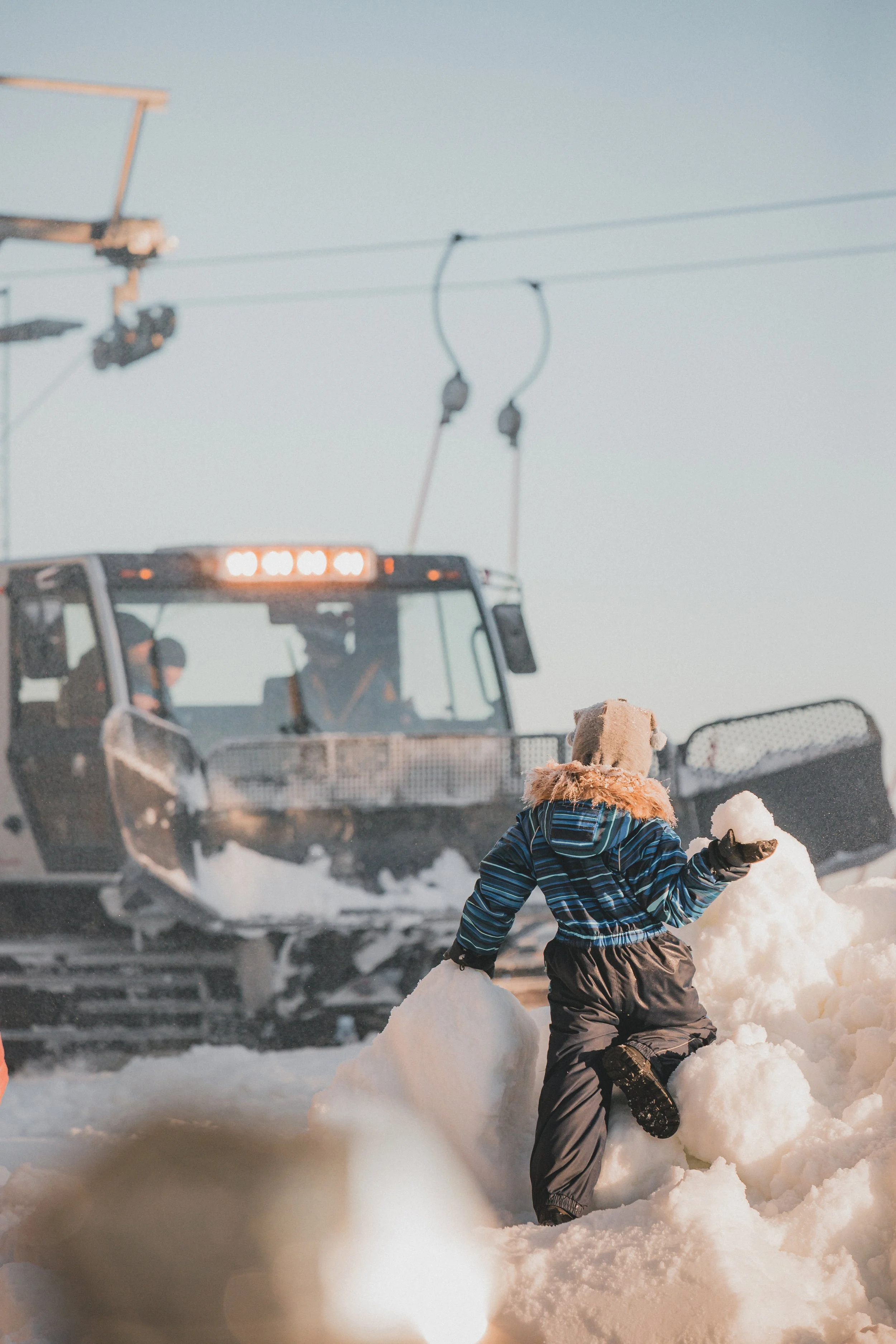 Child dressed in winter clothing and a bear hat, shoveling snow on a snowy landscape near a ski lift, with a snow groomer vehicle in the background.