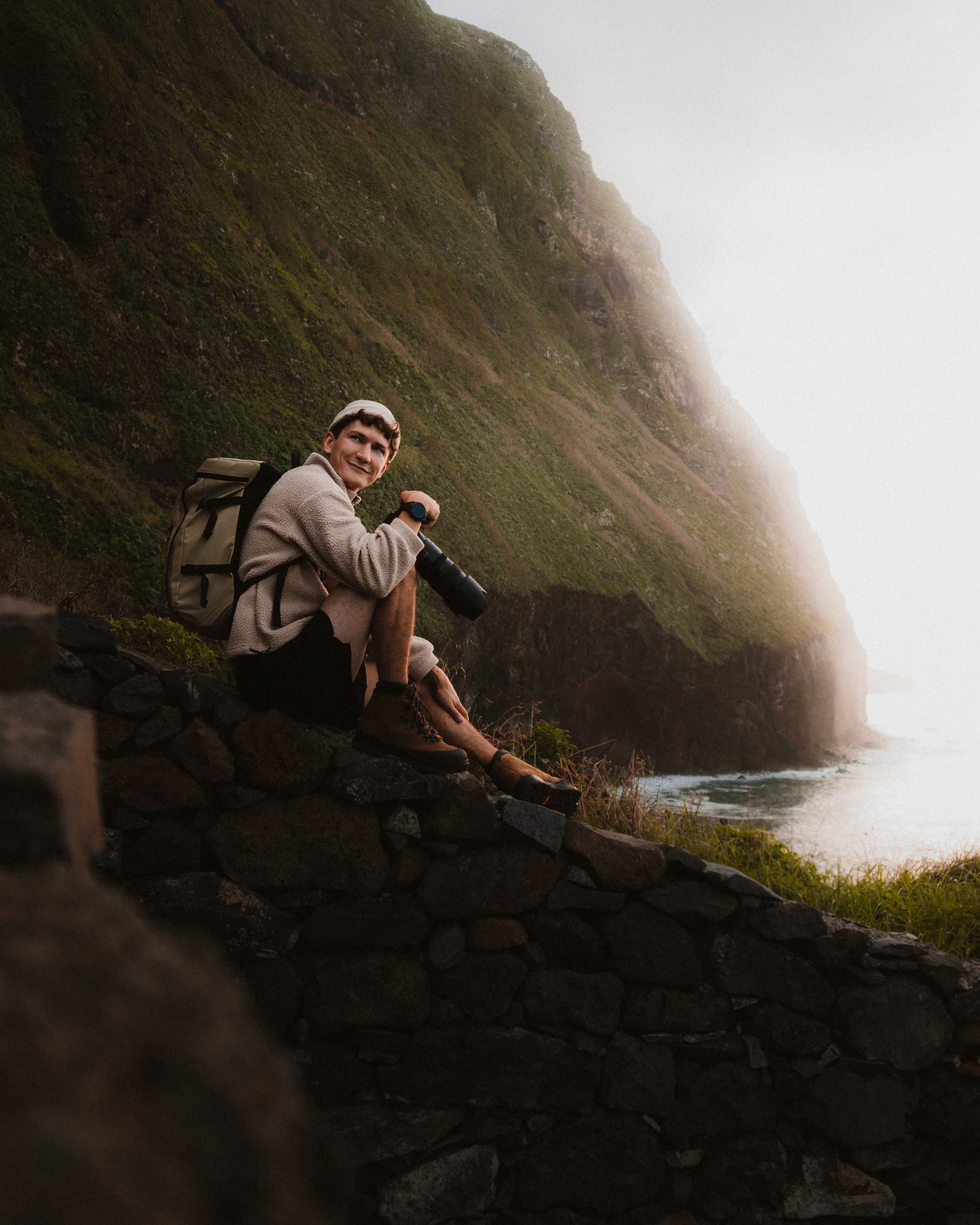 Photogrpaher sitting on an edge with camera in hand. Behind him are beutiful cliffs with golden light