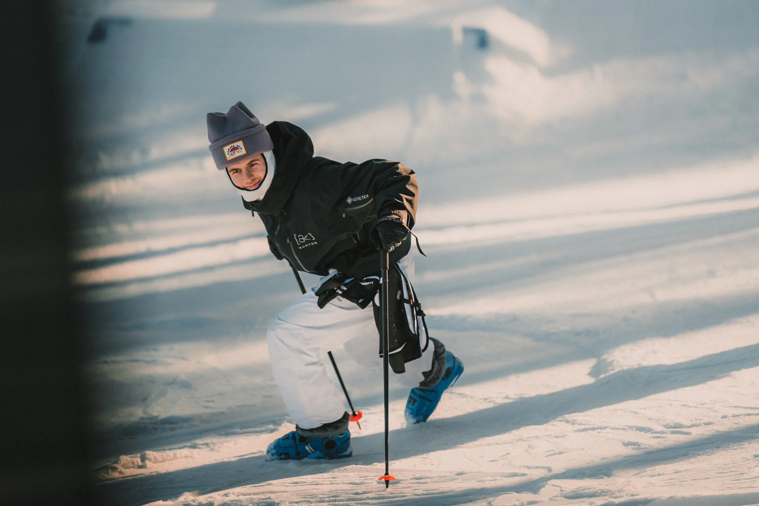 A person in skiing gear crouching on snow, smiling, holding ski poles, with ski boots on snow, wearing a gray beanie and black winter jacket.