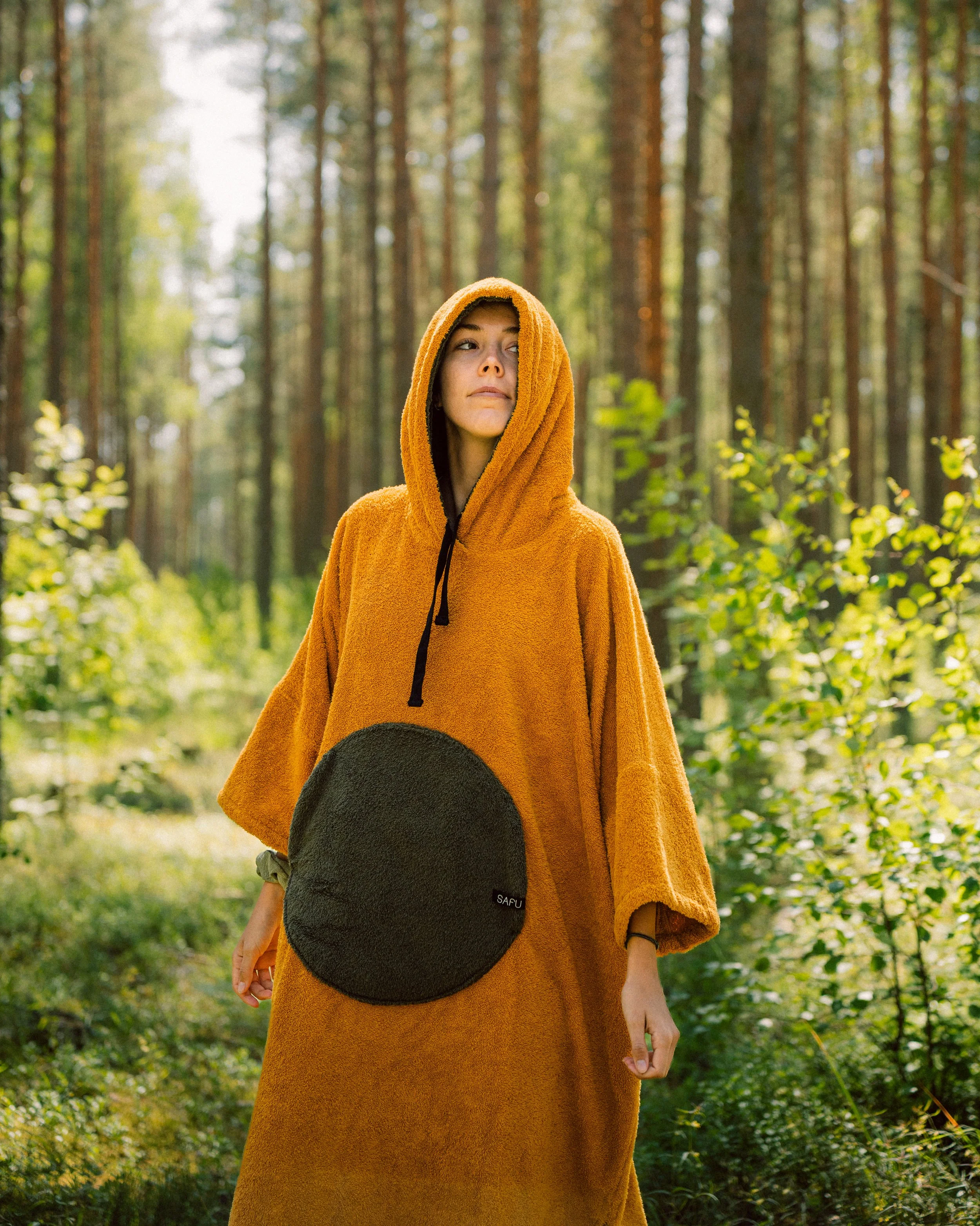 Person in a yellow towel poncho with a hood, standing in a forest with tall trees and green foliage.