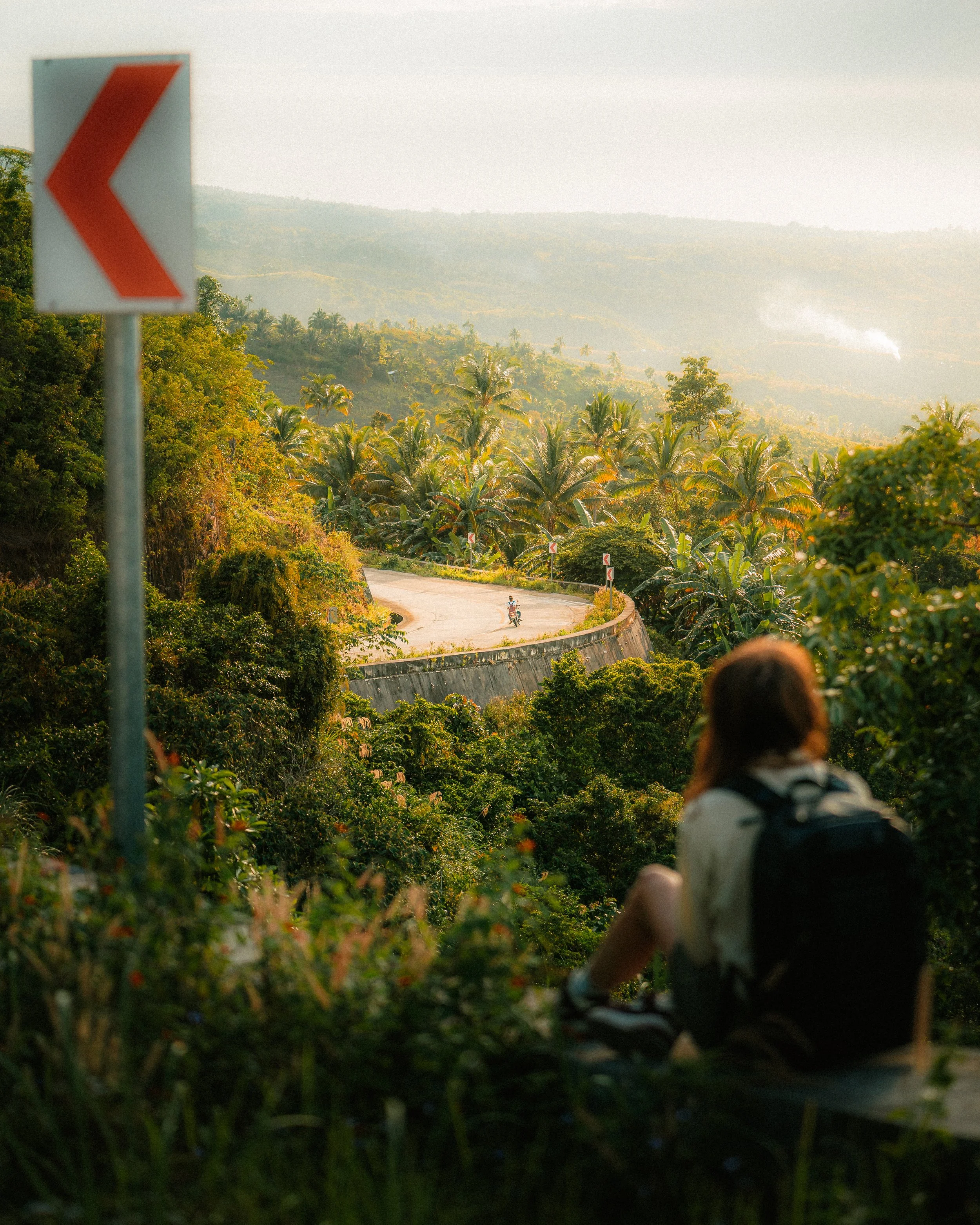 A golden light photo where a person is overlooking a tropical mountain landscape and a curvy road where there is a driver on a motorbike