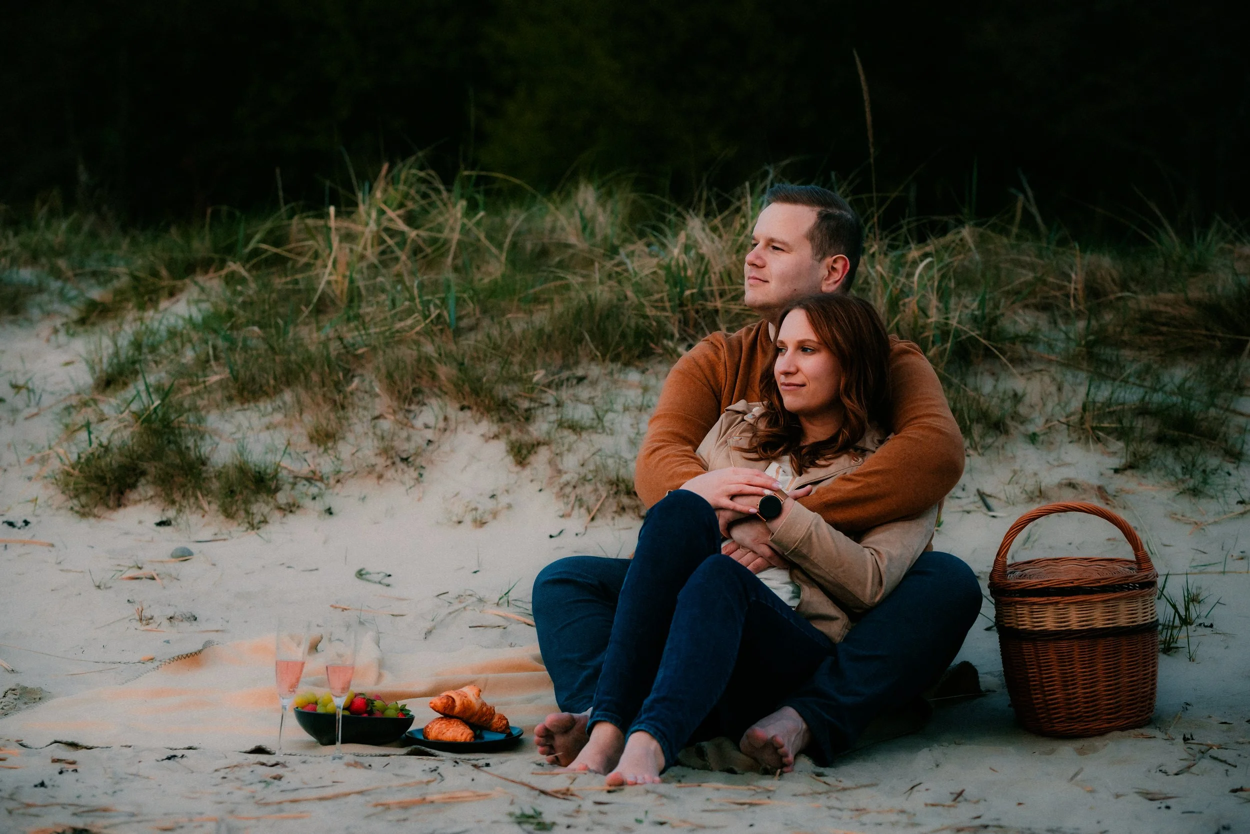 A couple sitting on a blanket on the beach with a picnic basket, snack, and drinks, enjoying the sunset.
