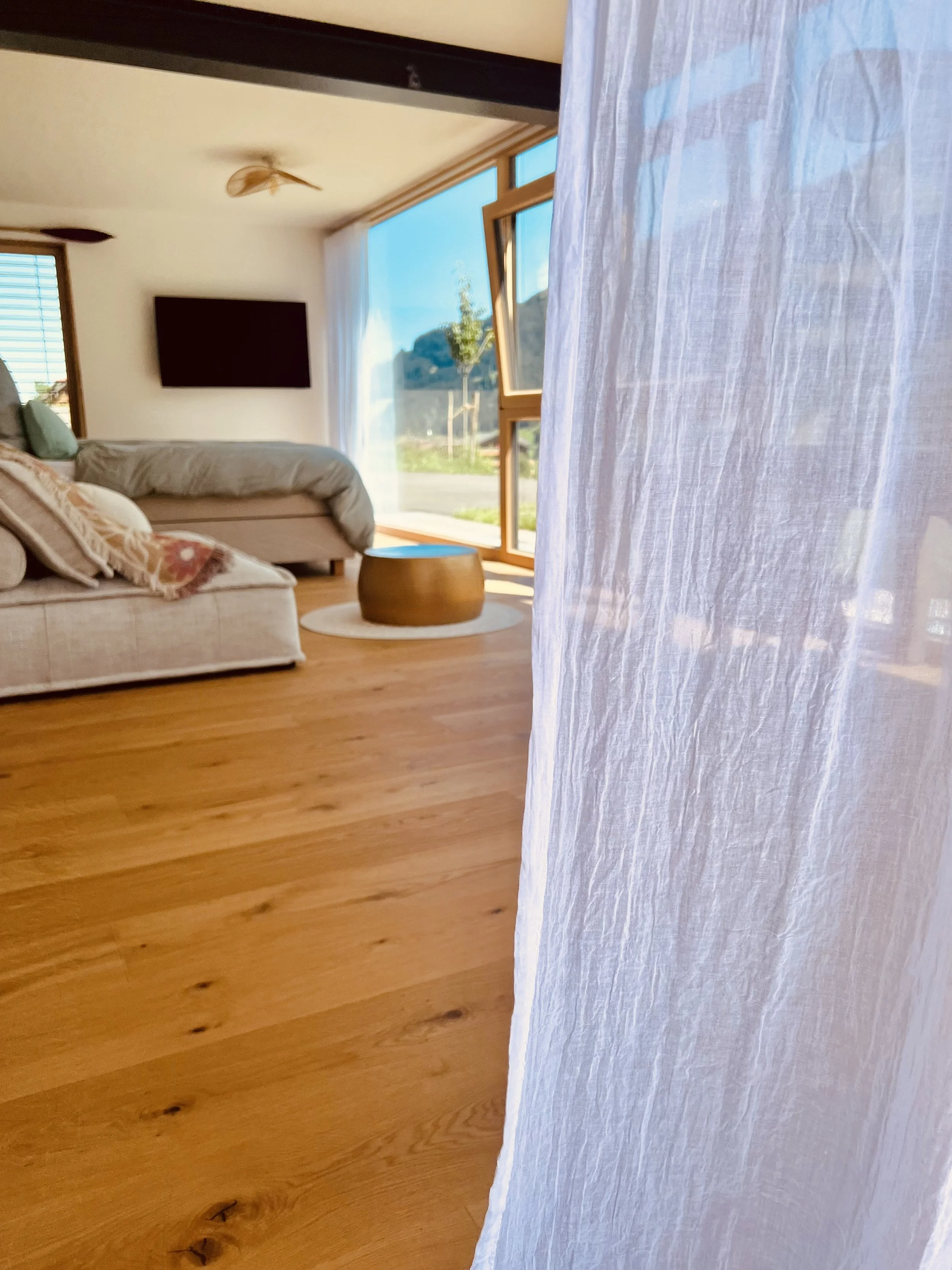 Ein gemütliches Wohnzimmer mit großen Fenstern, Holzboden, Sofa mit Kissen, Fernseher an der Wand und einem kleinen runden Tisch.