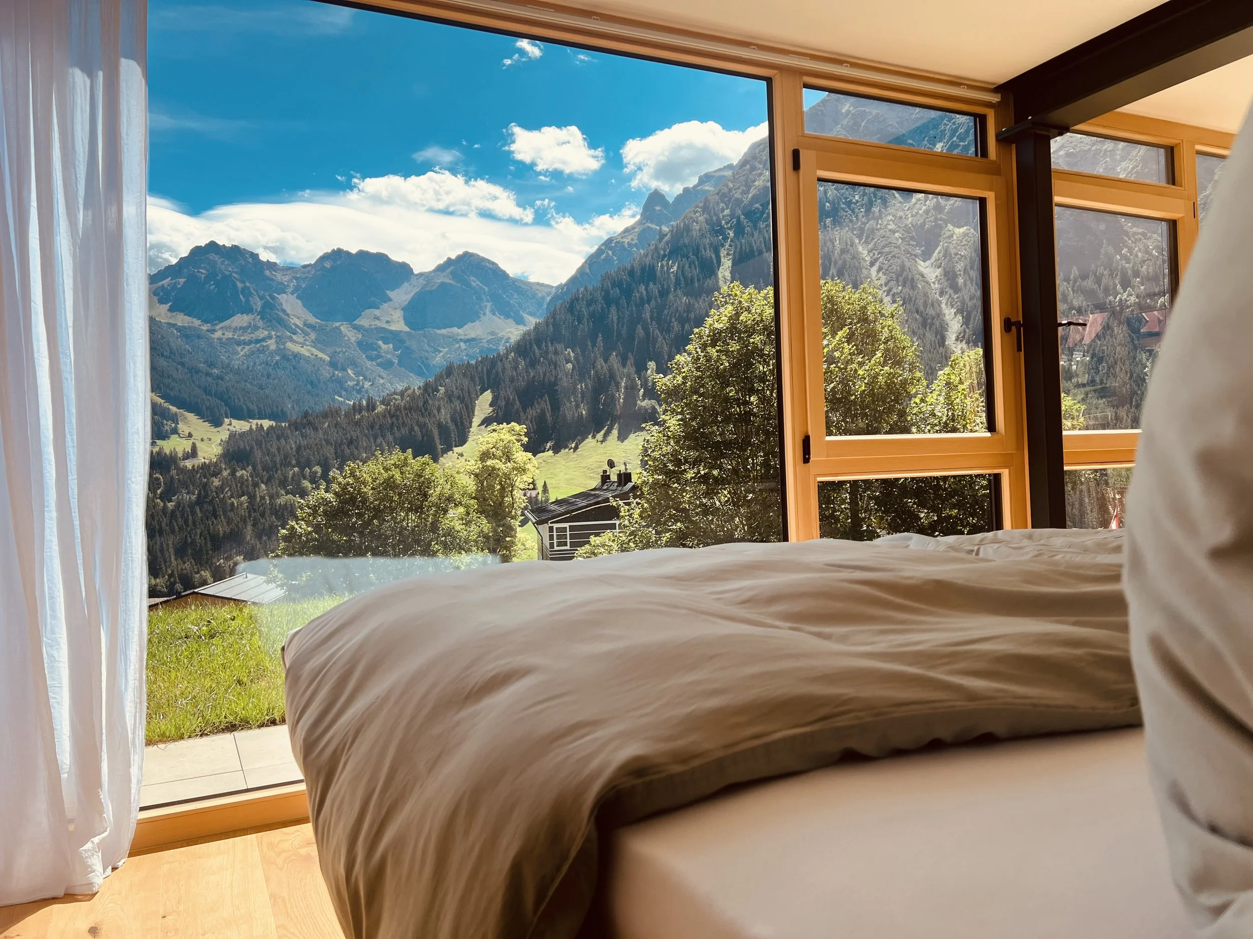 Blick aus einem offenen Fenster in ein Schlafzimmer mit Bergkulisse, grünen Hügeln und Wäldern bei sonnigem Wetter.
