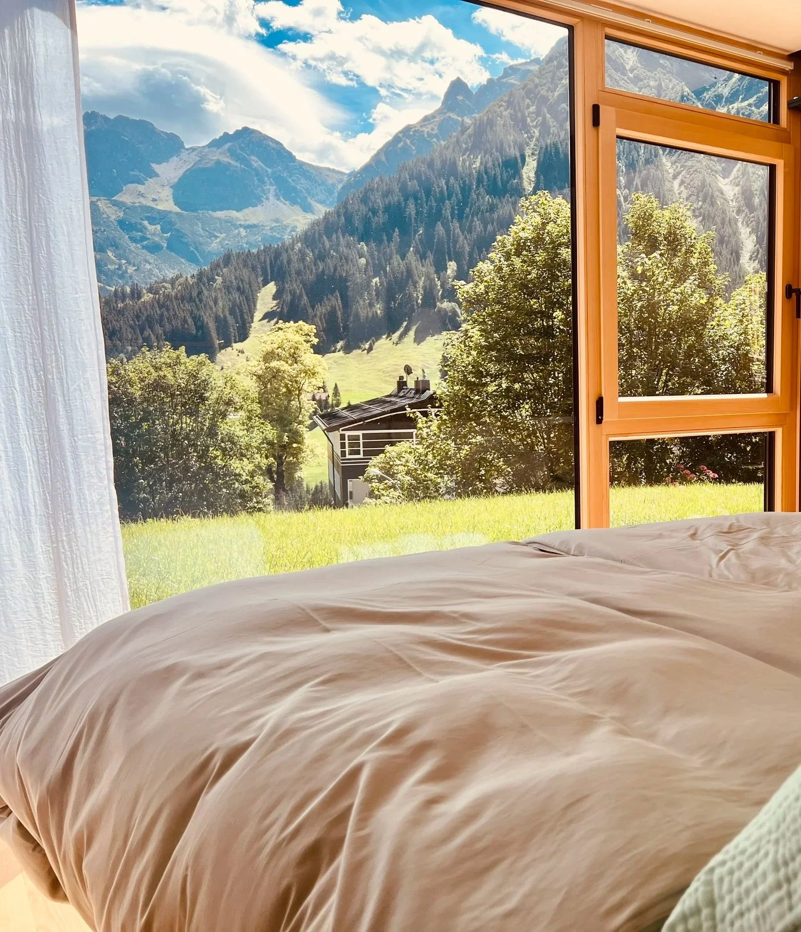 Blick aus einem Zimmer auf eine grüne Wiese und Berge im Hintergrund, offene Fenster, Bett im Vordergrund.