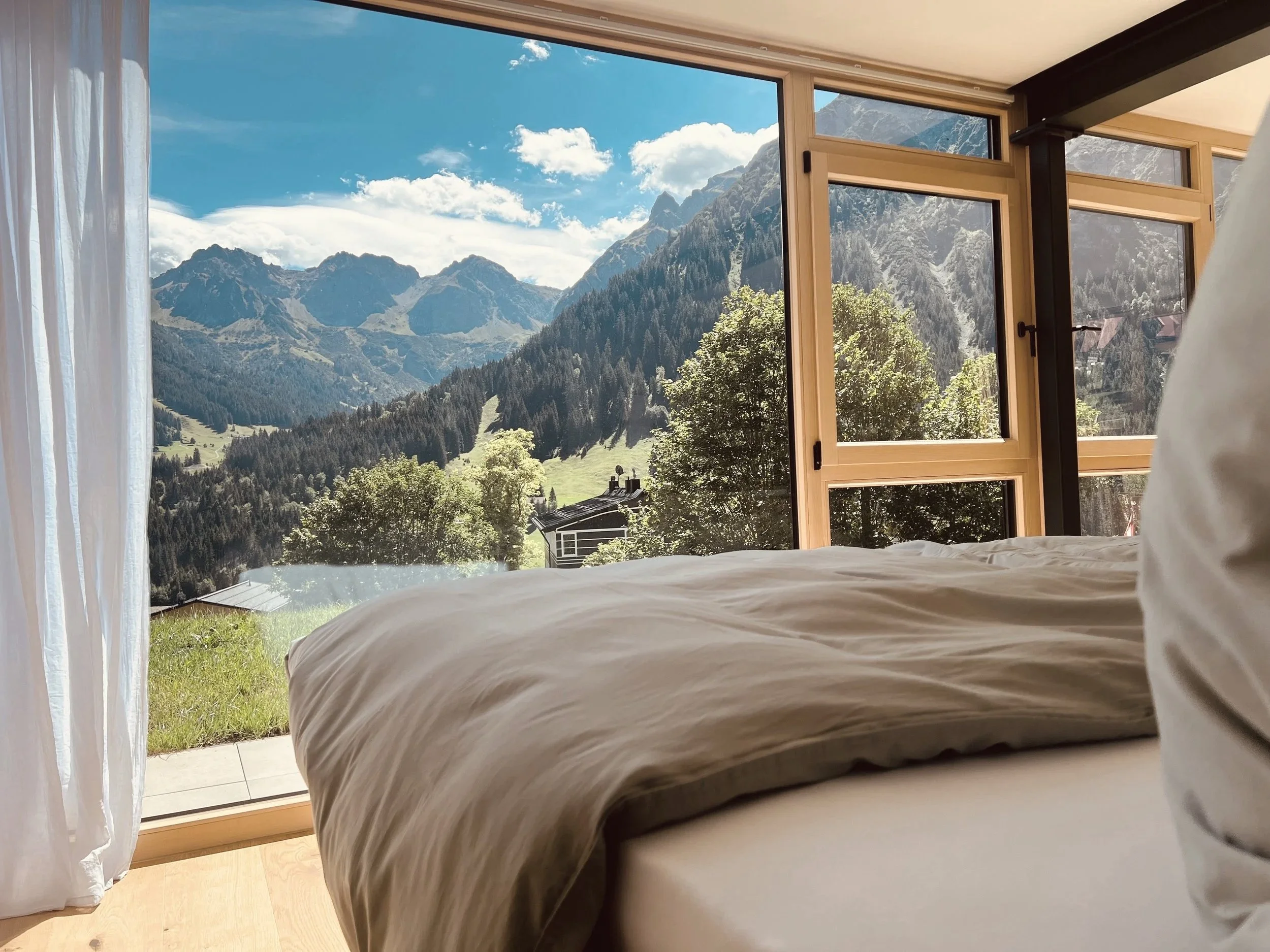 Blick aus einem Schlafzimmer mit offenen Fenstern auf Berge, Bäume und Himmel.