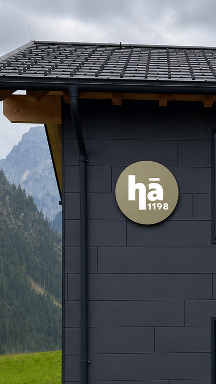 Außenansicht eines modernen Gebäudes mit schwarzen Fassaden, Hüttenstil, mit einem runden Logo mit der Beschriftung 'hä 1198', umgeben von einer alpinen Landschaft mit Bergen und Wäldern.