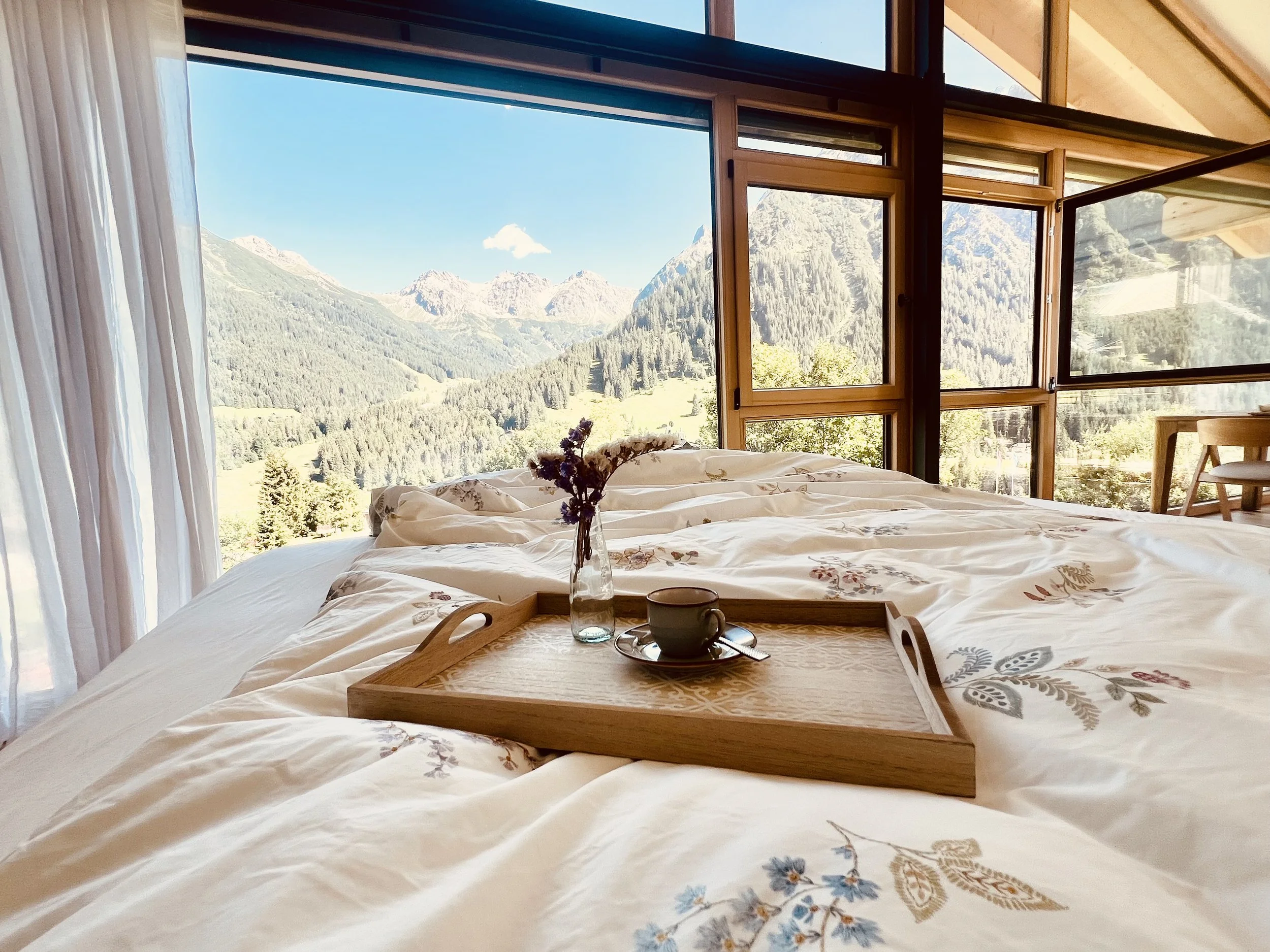Blick auf ein Schlafzimmer mit großen Fenstern, die eine Berglandschaft mit Bergen, Wäldern und blauen Himmel zeigen.