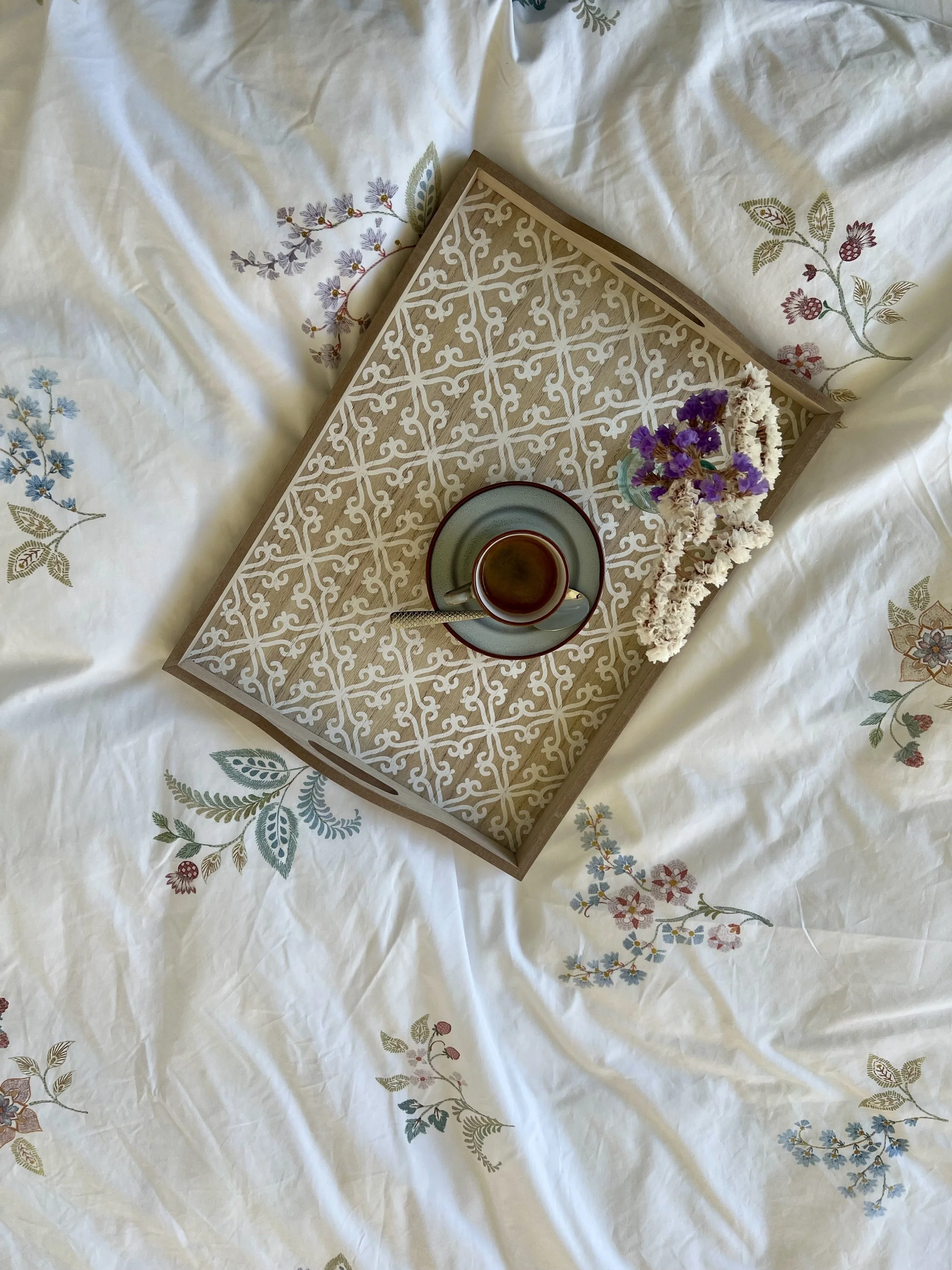 Ein Tablett mit einer Tasse Tee, einer kleinen Vase mit Lavendelblumen und einem Kranz aus getrockneten Blumen auf einem bedruckten Bettwäsche