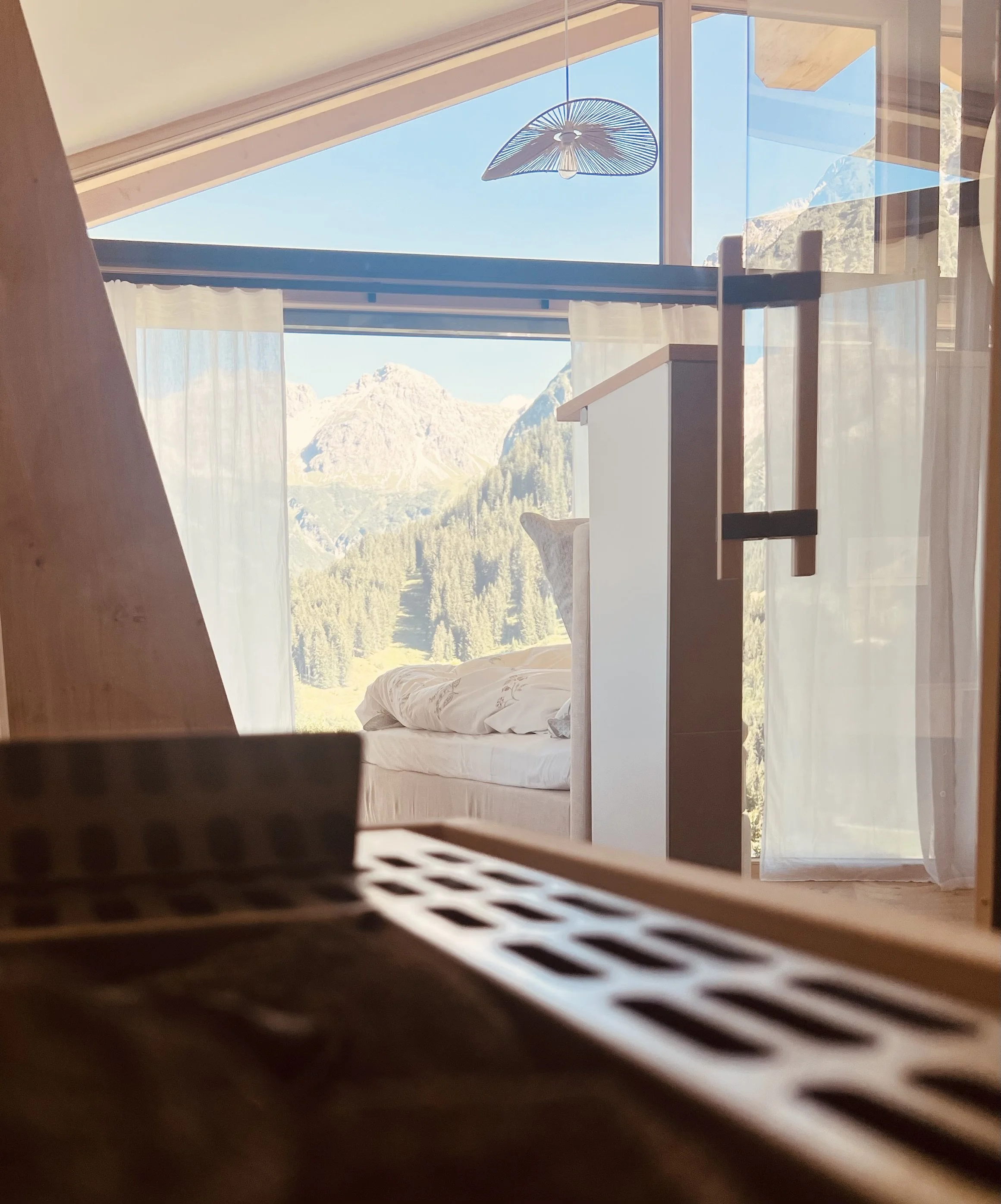 Helles Zimmer mit großen Fenstern, Blick auf Berge, Bett mit weißen Bettdecke, Vorhänge, moderner Kronleuchter.