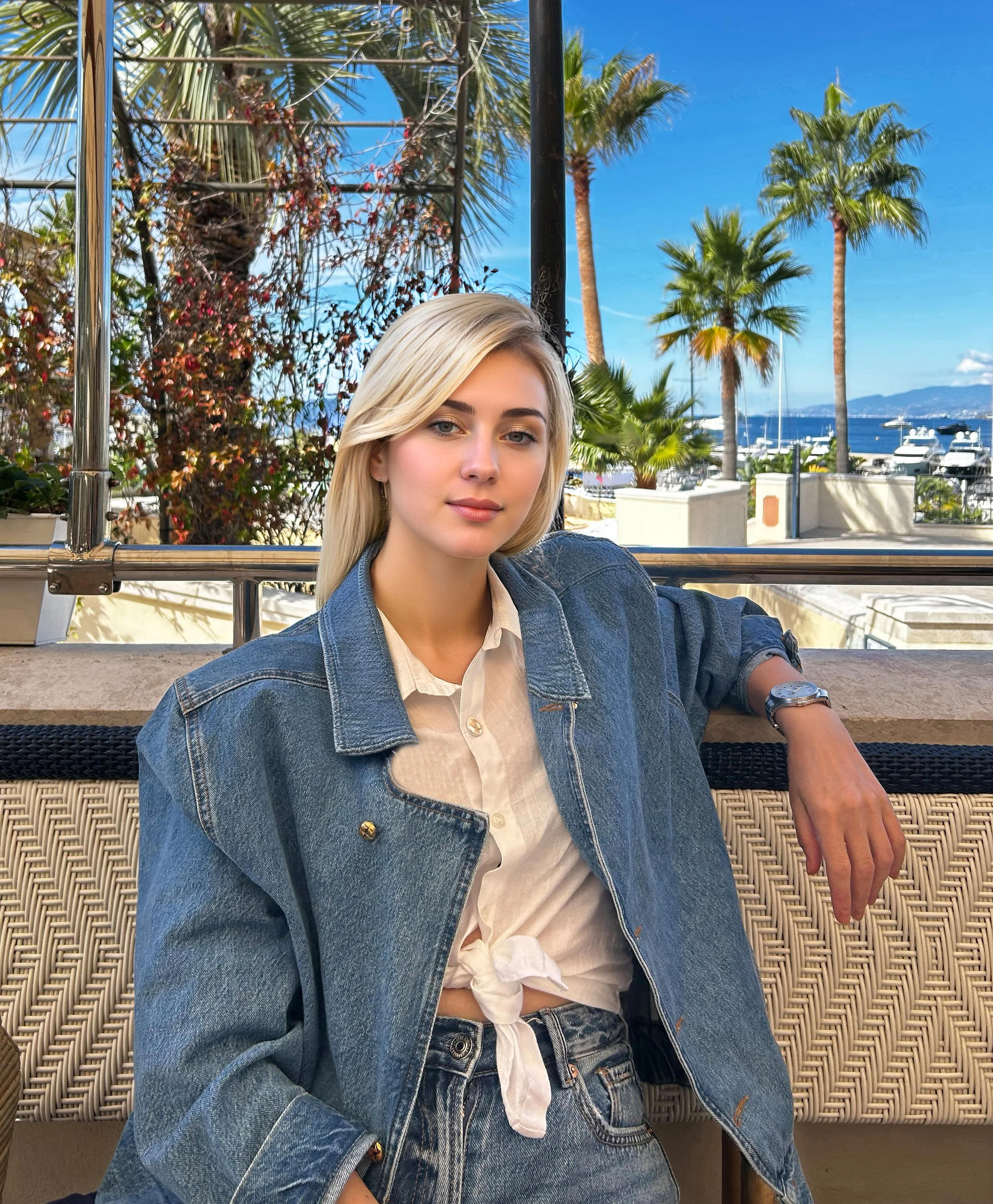 A young woman with blonde hair and fair skin, wearing a denim jacket and cream-colored top. She is sitting outdoors with palm trees, boats, and a body of water in the background, under a clear blue sky.