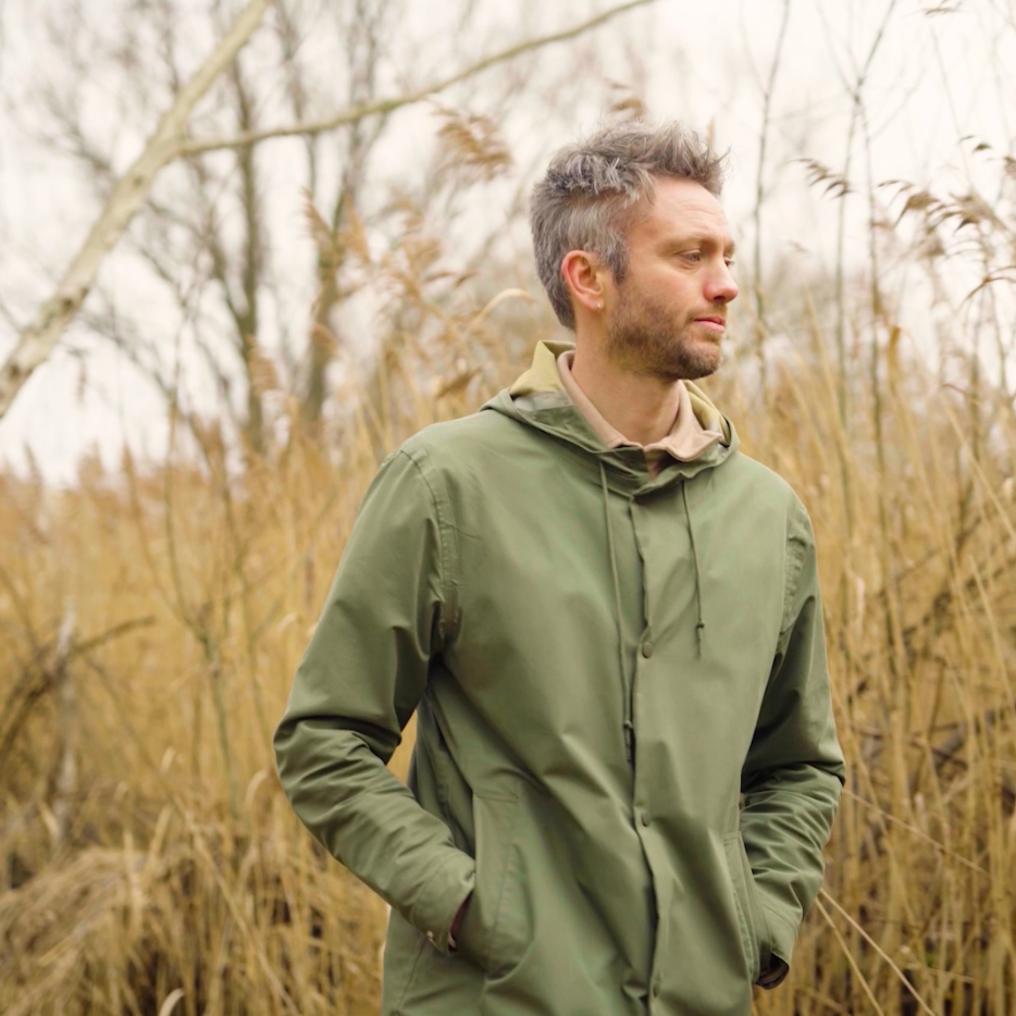 Man in groene jas staat in een veld met bruine vegetatie.