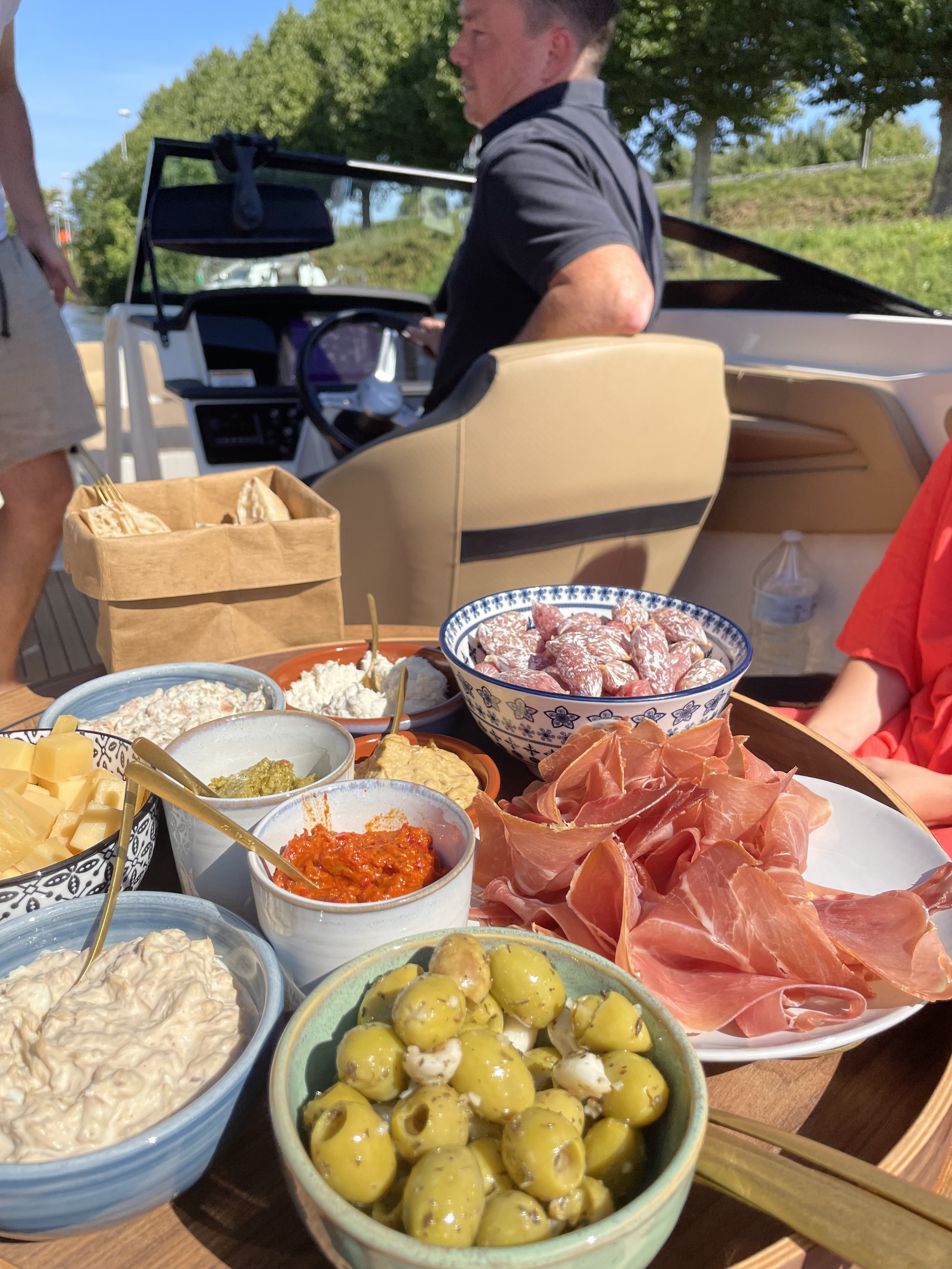 Een tafel met diverse hapjes waaronder olijven, ham, kaas, tapenade en rauwe vleeswaren, met een boot op de achtergrond en mensen op een boot.