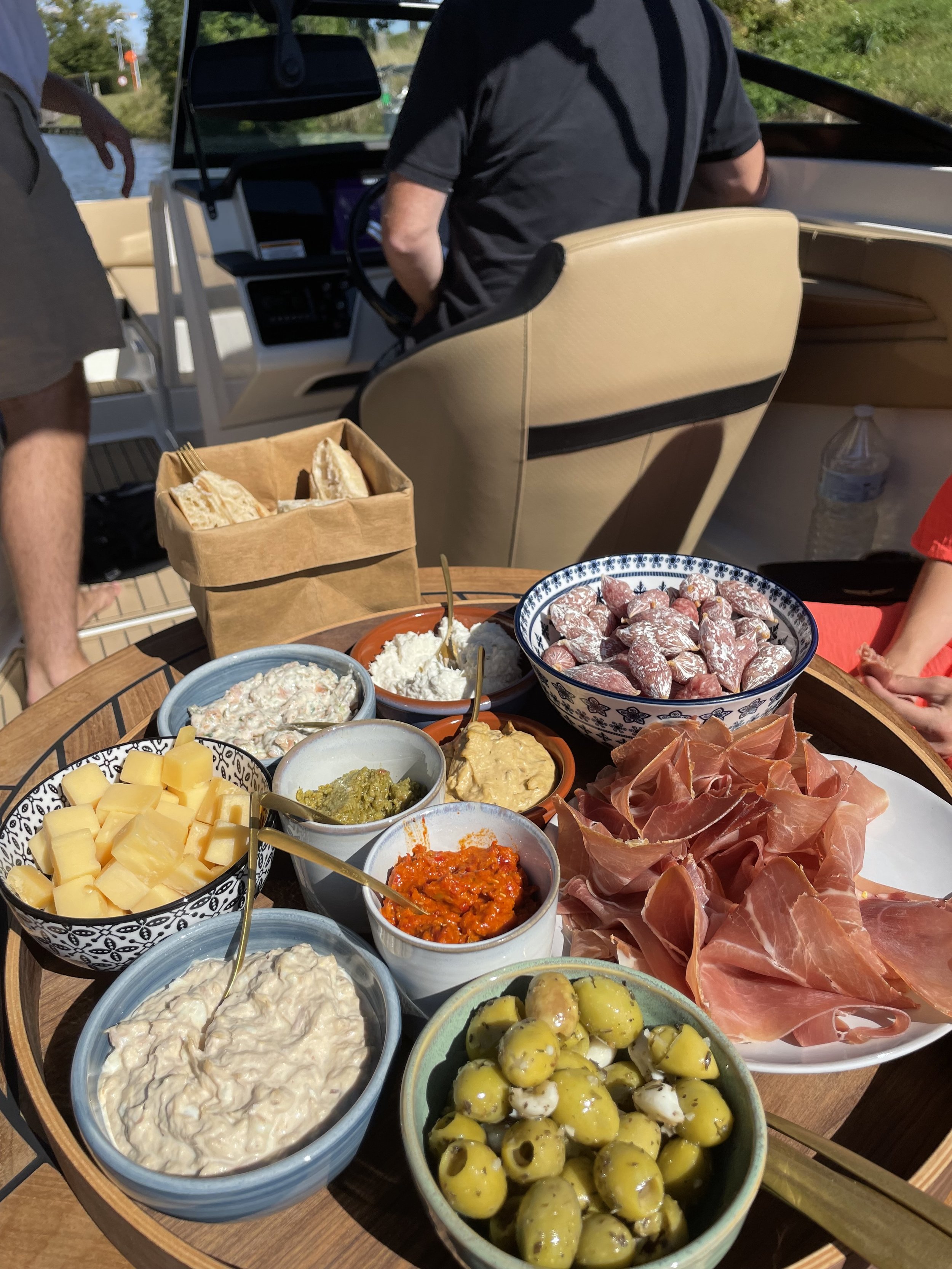 Een tafel gevuld met diverse hapjes zoals kaas, olijven, ham, tapenade en andere dips, met achtergrond van een boot met mensen.